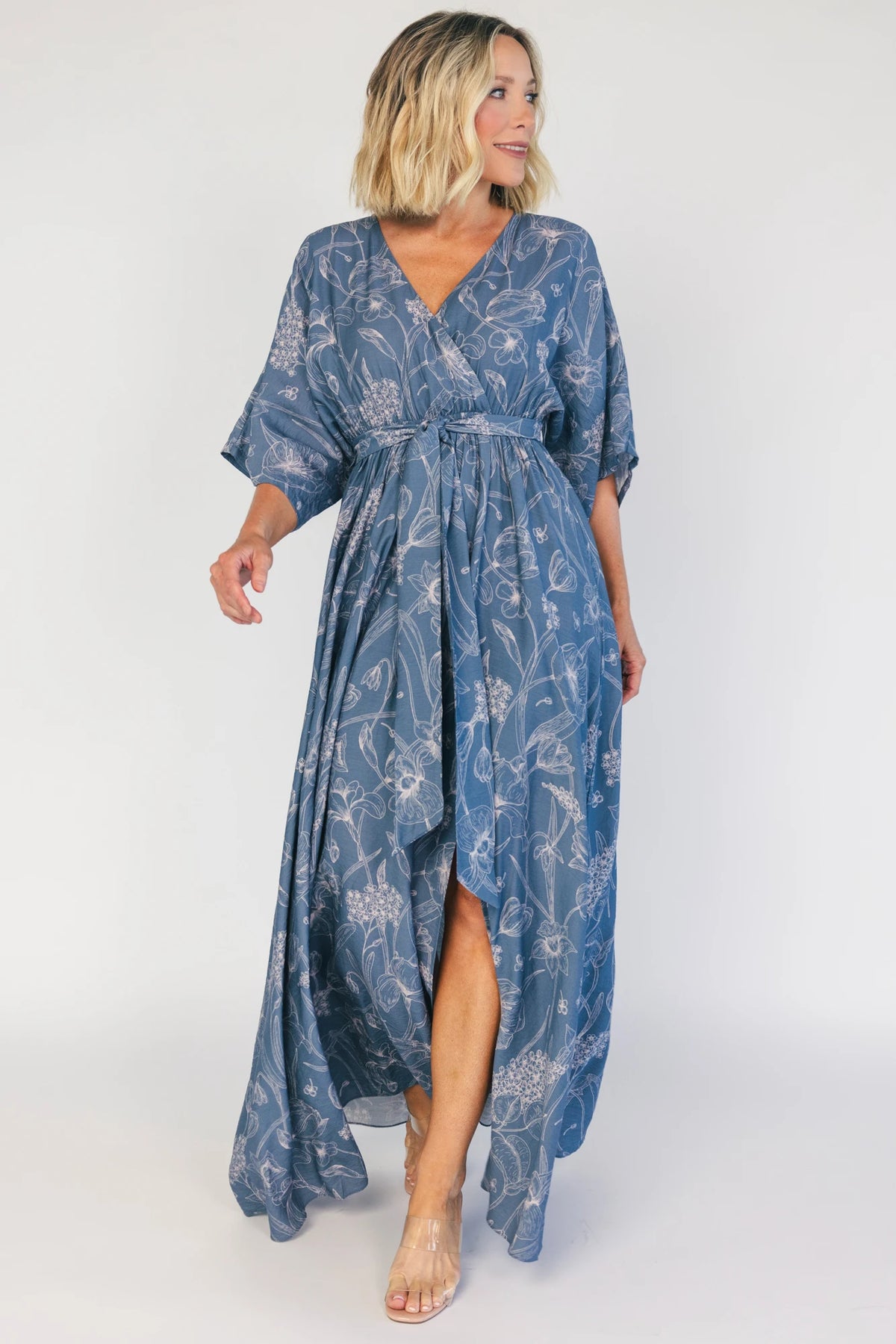 Model wearing a navy botanical-print maxi dress with a surplice V-neckline, kimono sleeves, an elastic waist with a removable sash tie, and a flowing full-length skirt featuring a side slit.