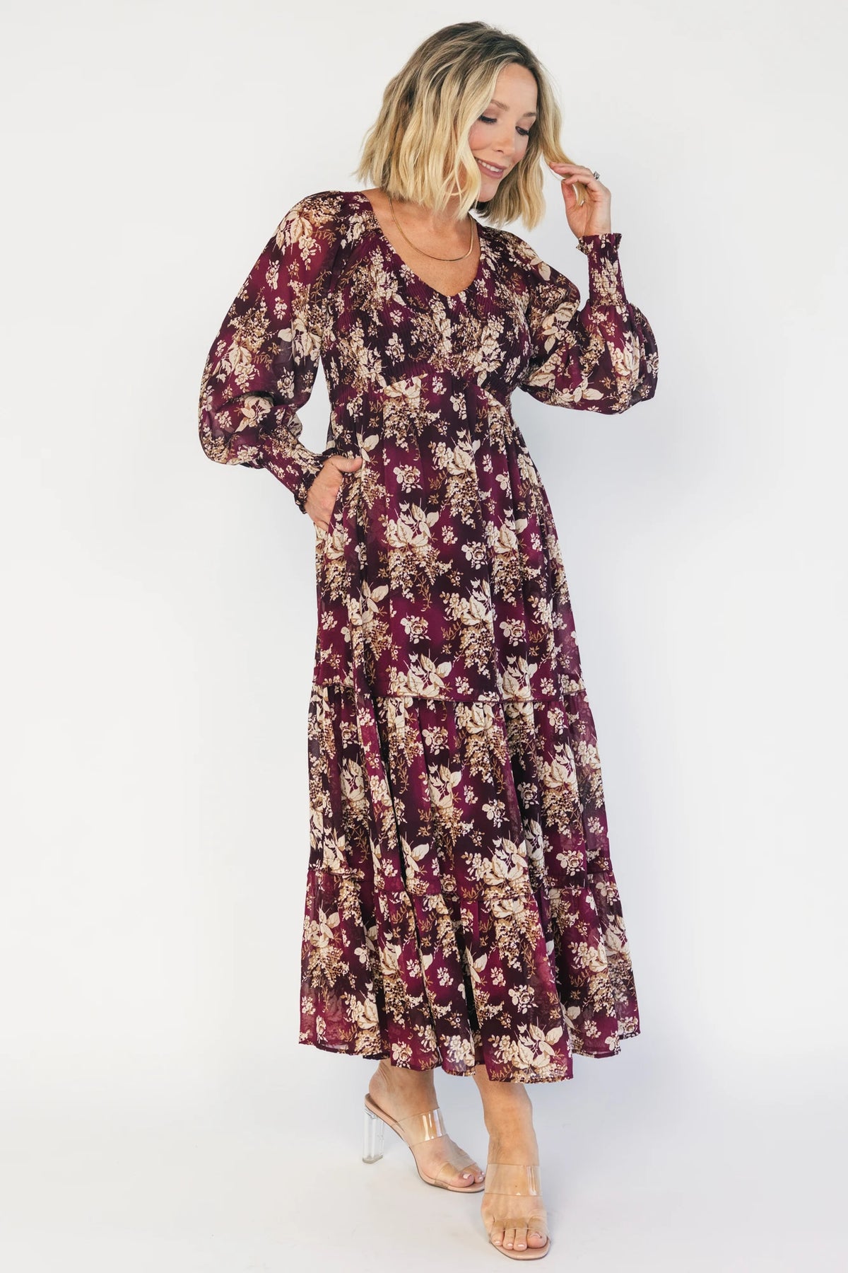 Model wearing a burgundy floral maxi dress with a V-neckline, smocked bodice, and long sheer sleeves with smocked ruffle cuffs.