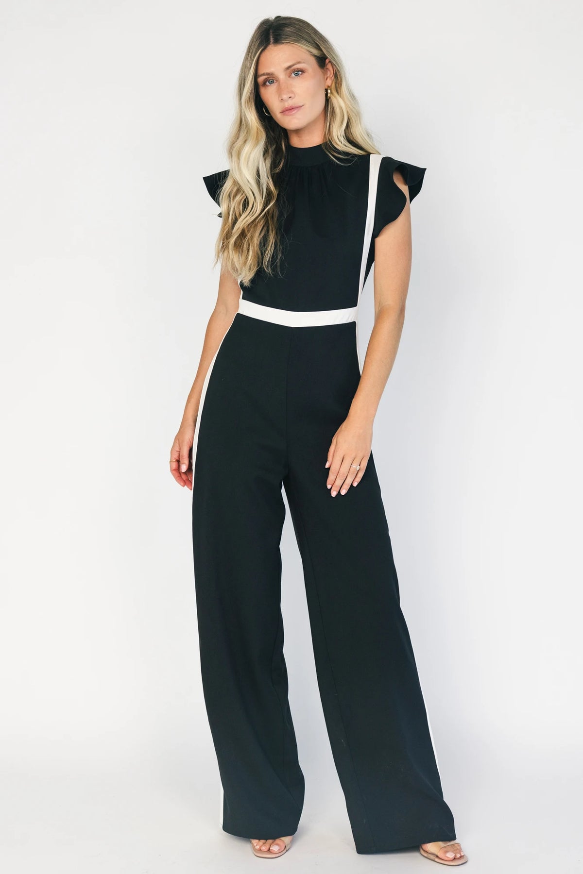 Model wearing the Lumina Ruffle Jumpsuit in Black + Ivory featuring a sleeveless ruffle-trim bodice, defined waist, and flowing wide-leg pants.