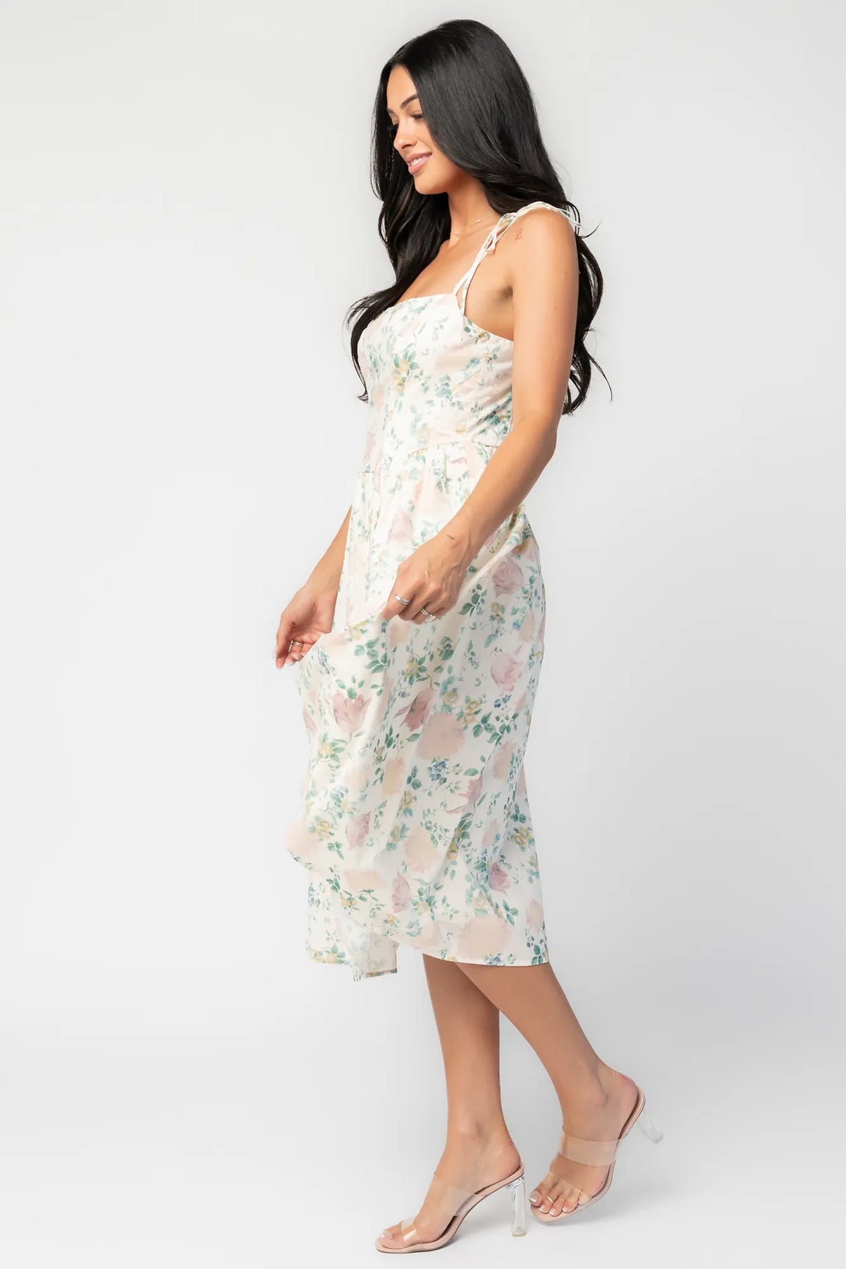 Side image of model wearing a cream floral midi dress with a square neckline, adjustable shoulder tie straps, fitted bodice, and flowing skirt.