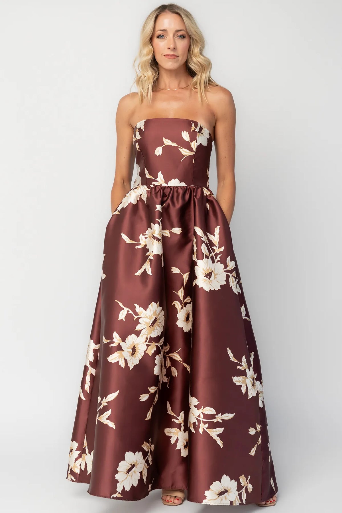 Model wearing a strapless maxi gown with a mahogany floral print and a fitted bodice. A rosette detail accents the neckline, while the flowing floor-length skirt creates elegant movement.