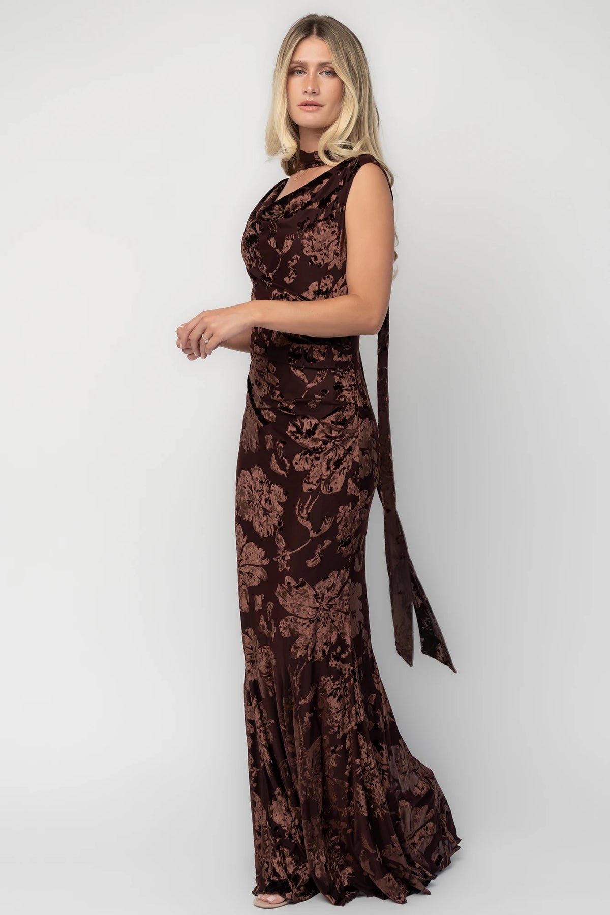 Model wearing a brown velvet burnout floral maxi dress with a draped cowl neckline, scarf detail, side ruching, and a flowing mermaid silhouette.