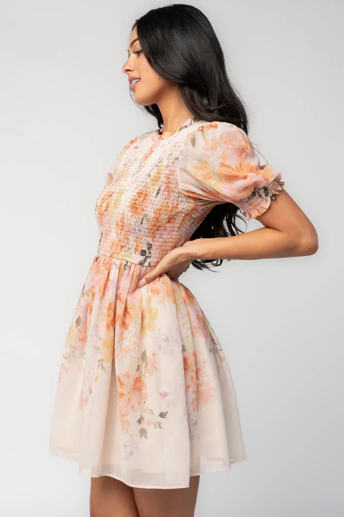 Model wearing an ivory floral short dress with a fully smocked bodice, ruffled round neckline, and short puff sleeves.