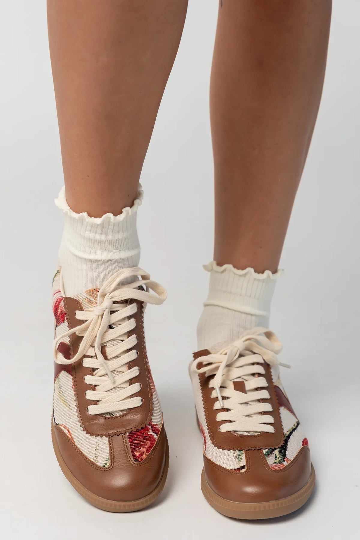 Pair of low-top sneakers featuring cognac faux leather overlays, floral tapestry panels with rose motifs, and cream laces. The vintage-inspired design combines mixed textures and warm tones for a casual yet elevated everyday look.