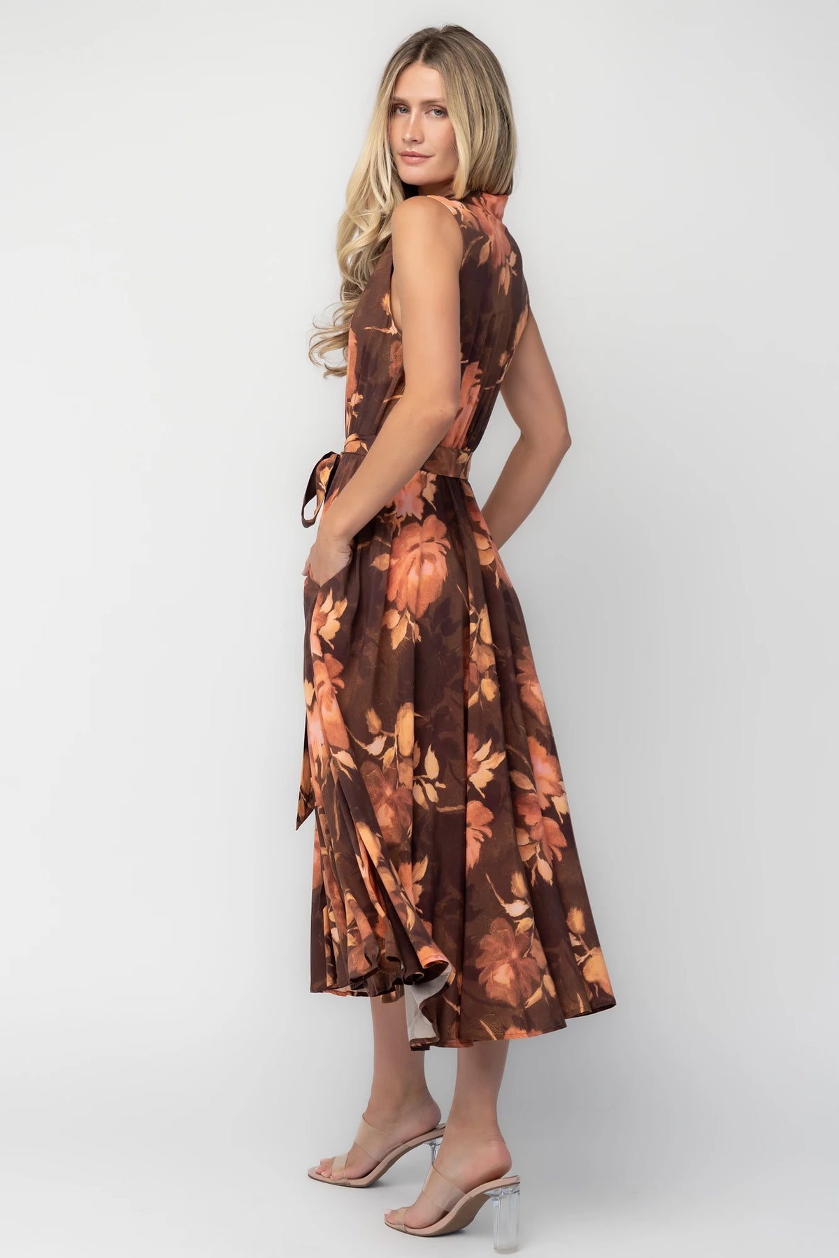 Model wearing the Tess Midi Dress in Brown + Rust featuring a collared neckline, functional button-front bodice, sleeveless design, removable waist tie, elastic back waist, hidden side pockets, and a flowing midi skirt with a brown background and warm rust floral print.