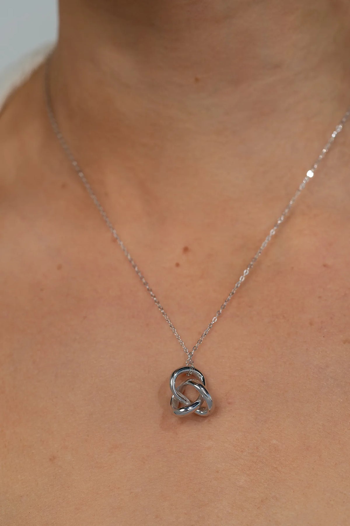 Model wearing a delicate silver-tone necklace featuring an interlocking infinity knot pendant suspended from a fine chain. The minimalist design sits at the collarbone for an elegant, everyday look.