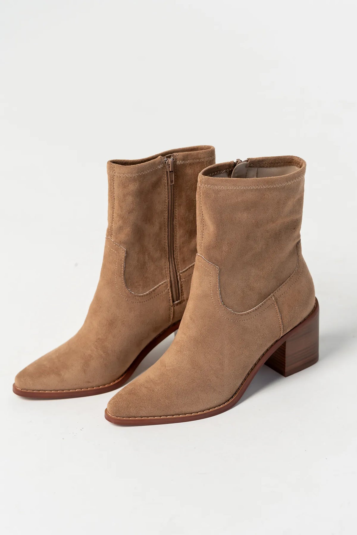 Pair of sand-colored ankle boots featuring a soft suede-like finish, pointed toe silhouette, and stacked block heel. The boots include a side zipper closure and subtle western-inspired seam detailing, creating a versatile look that pairs easily with denim, dresses, and seasonal outfits.