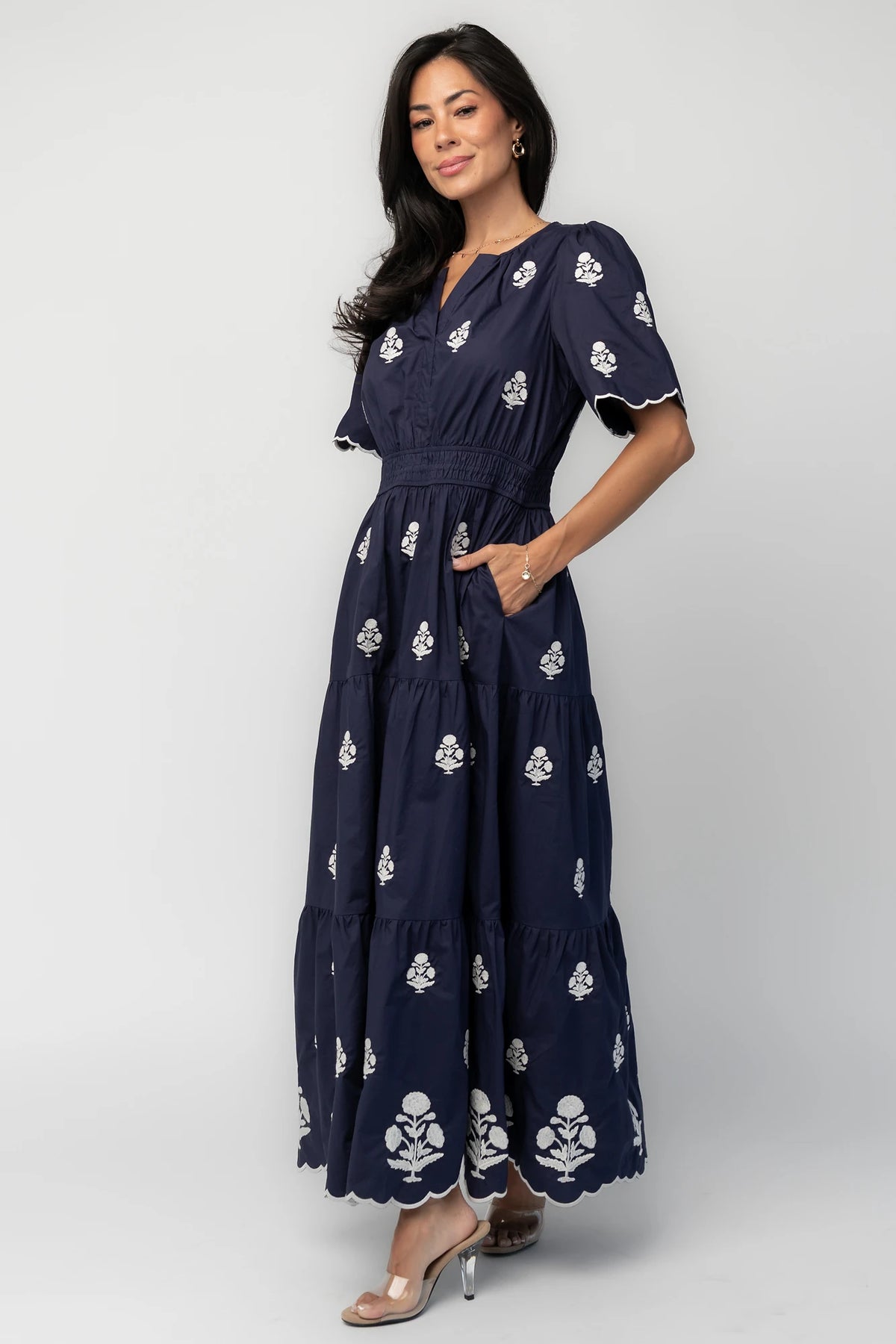 Model wearing the Delilah Embroidered Maxi Dress in navy featuring off-white floral embroidery, a split V-neckline, scalloped short sleeves, a smocked waistband, and a flowing tiered maxi skirt with a scalloped hem.