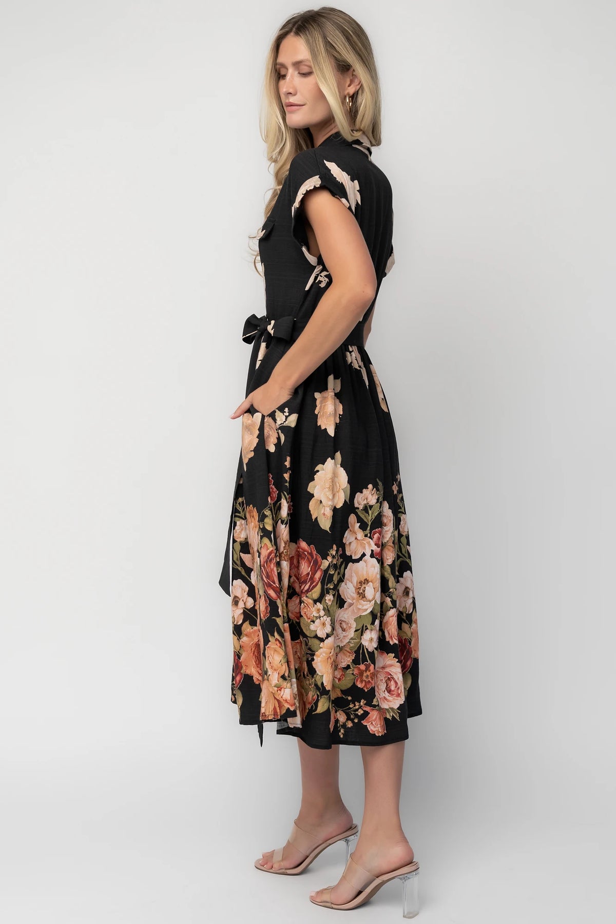 Model wearing a black floral midi dress with a collared neckline, functional button-front bodice, short sleeves, flap chest pockets, a removable waist tie, and a flowing midi-length skirt.
