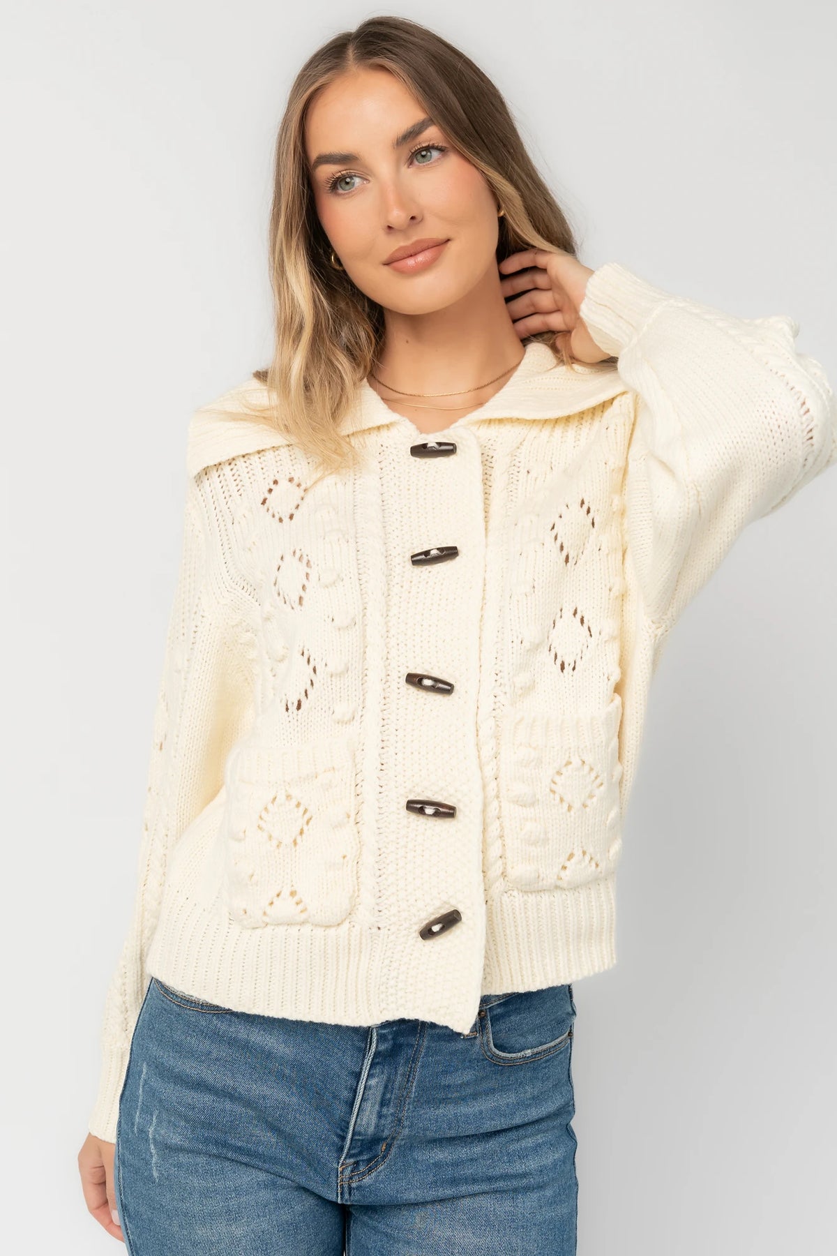 Cream cable knit sweater with a wide fold-over collar, toggle button front, long sleeves, bobble and pointelle knit detailing, ribbed trim, and a relaxed fit.