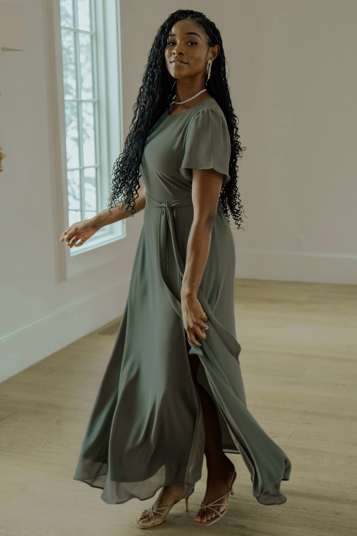 Studio detail shot of Naomi dark sage chiffon maxi dress focusing on hem sweep, left-leg slit, and the dress’s elegant movement.