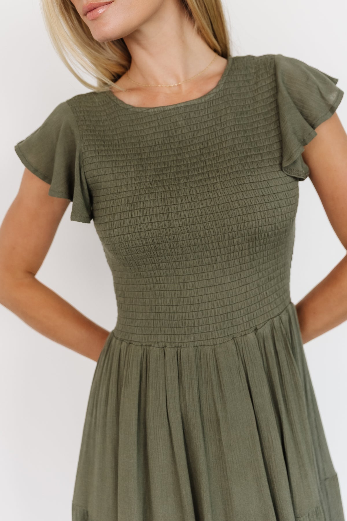 Model wearing Loretta Smocked Midi Dress in dark sage, angled side view emphasizing smocked waist, flutter cap sleeve, and flowy tiered hem.