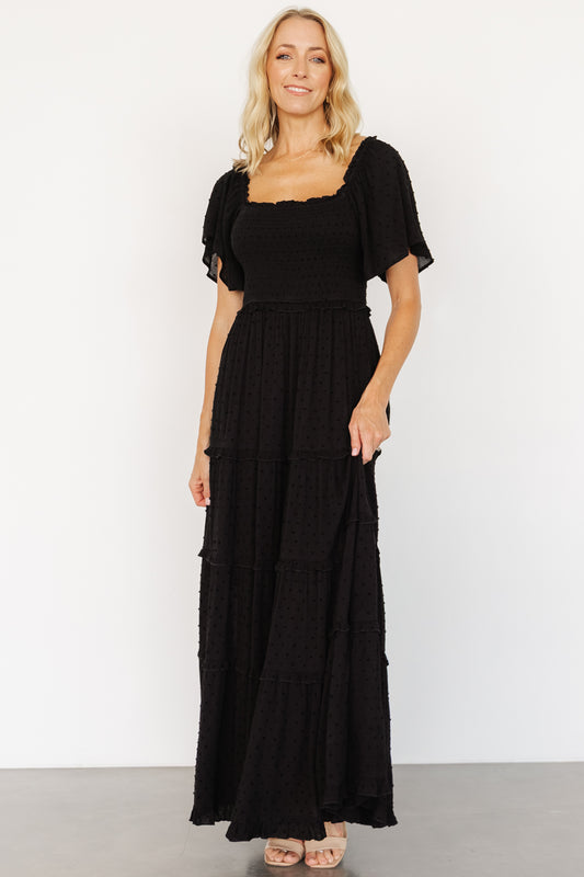 Three-quarter side view of black Lenora maxi dress on model; short flutter sleeves and ruffled elastic waist visible with soft drape of rayon fabric.