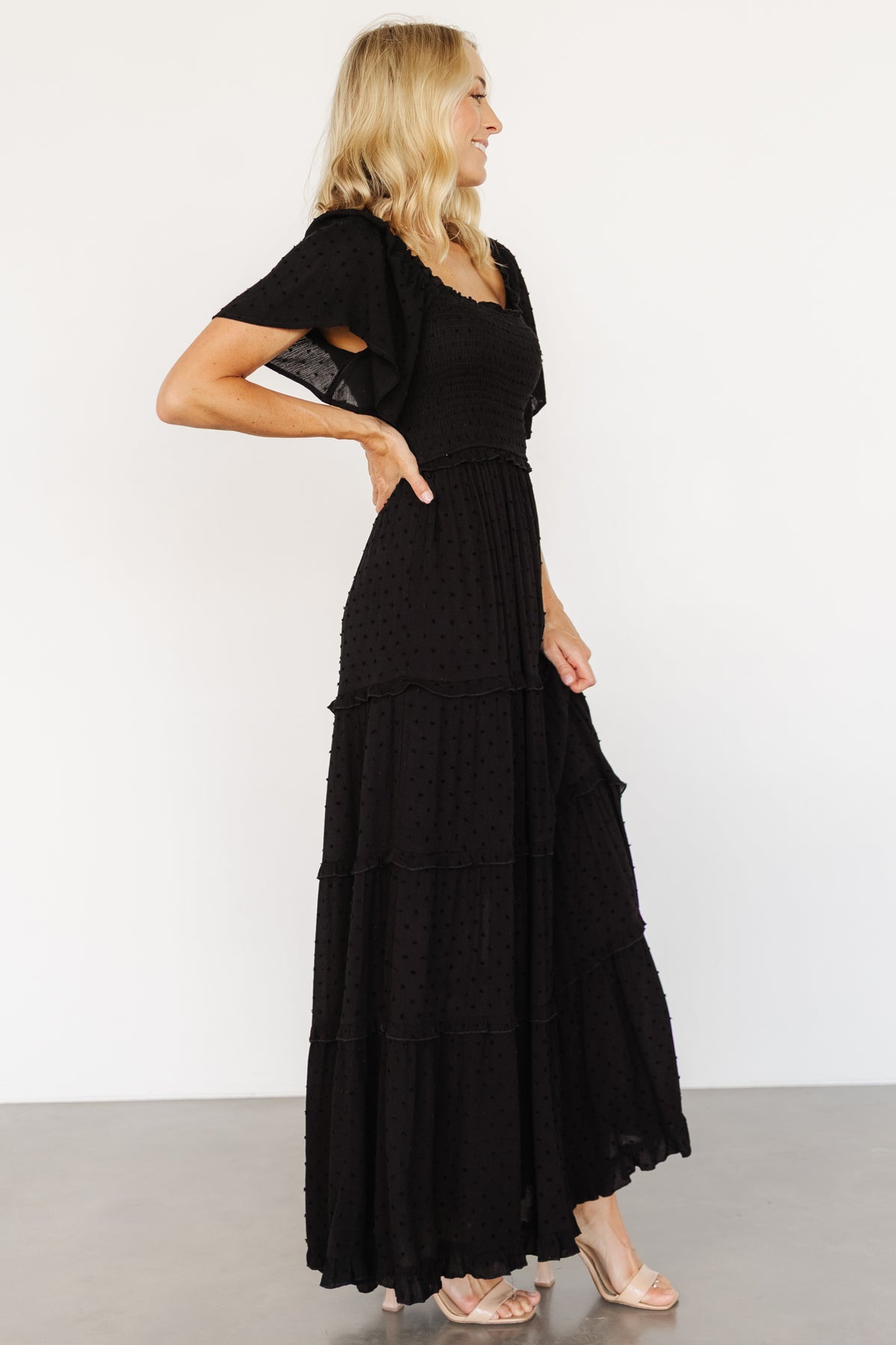 Model walking in Lenora black maxi dress, displaying natural fabric movement, tiered ruffle hem, and short flutter sleeves for a feminine finish.