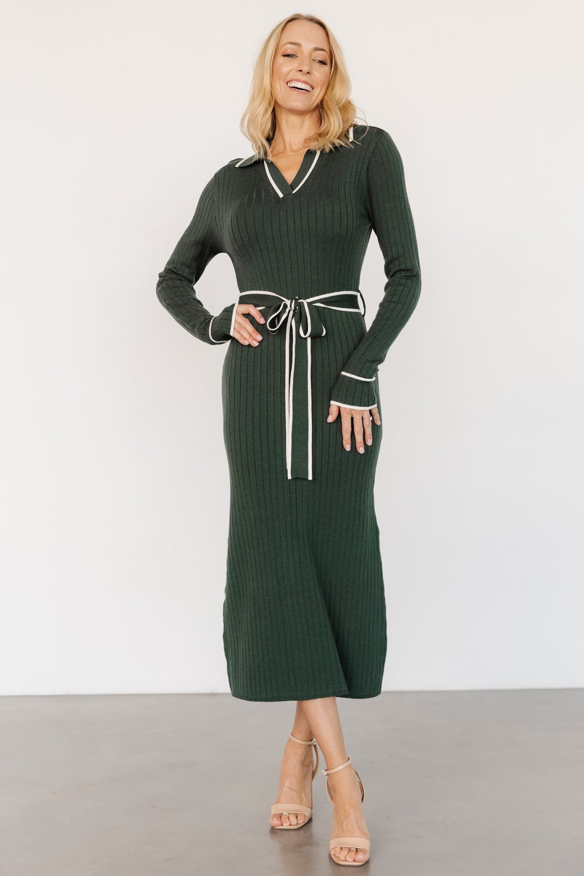 Front view of Lark ribbed sweater dress in green with ivory trim on a model, showing V-neck rugby-style collar and removable waist sash for styling.