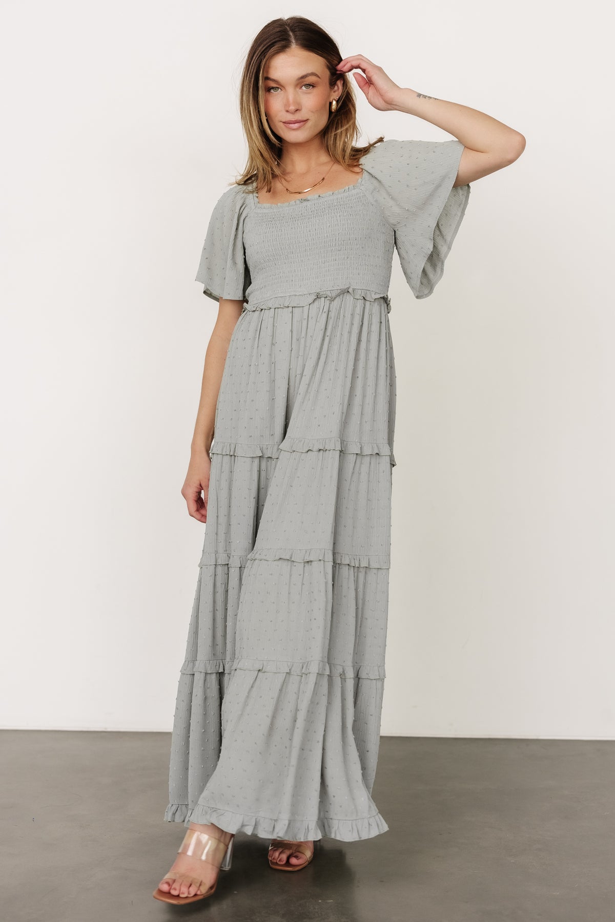 Model wearing Lenora maxi dress in sage, three-quarter view walking to show skirt movement, flutter sleeves, and dotted texture on the fabric.