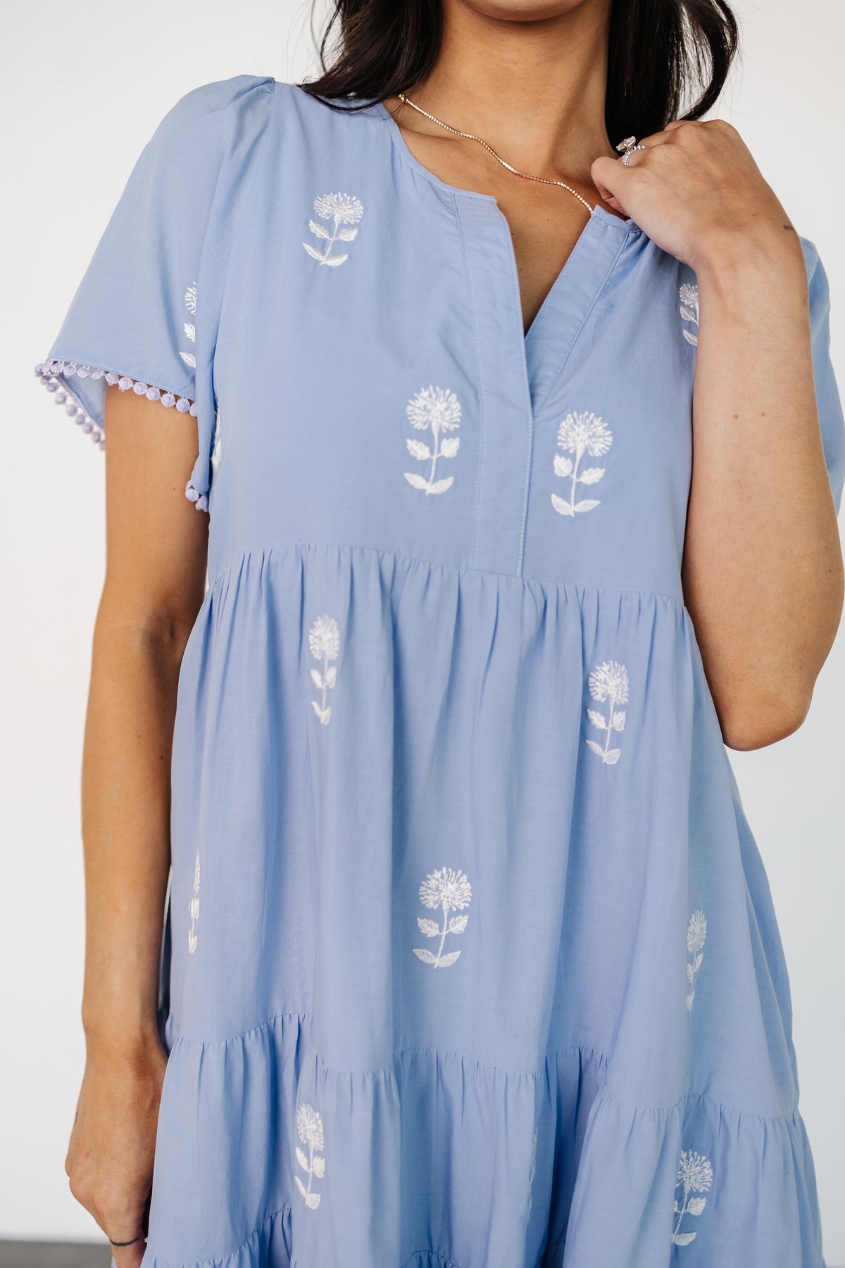 Model wearing ivory short dress with periwinkle embroidery, showing relaxed babydoll silhouette, short sleeves, and pocket placement.