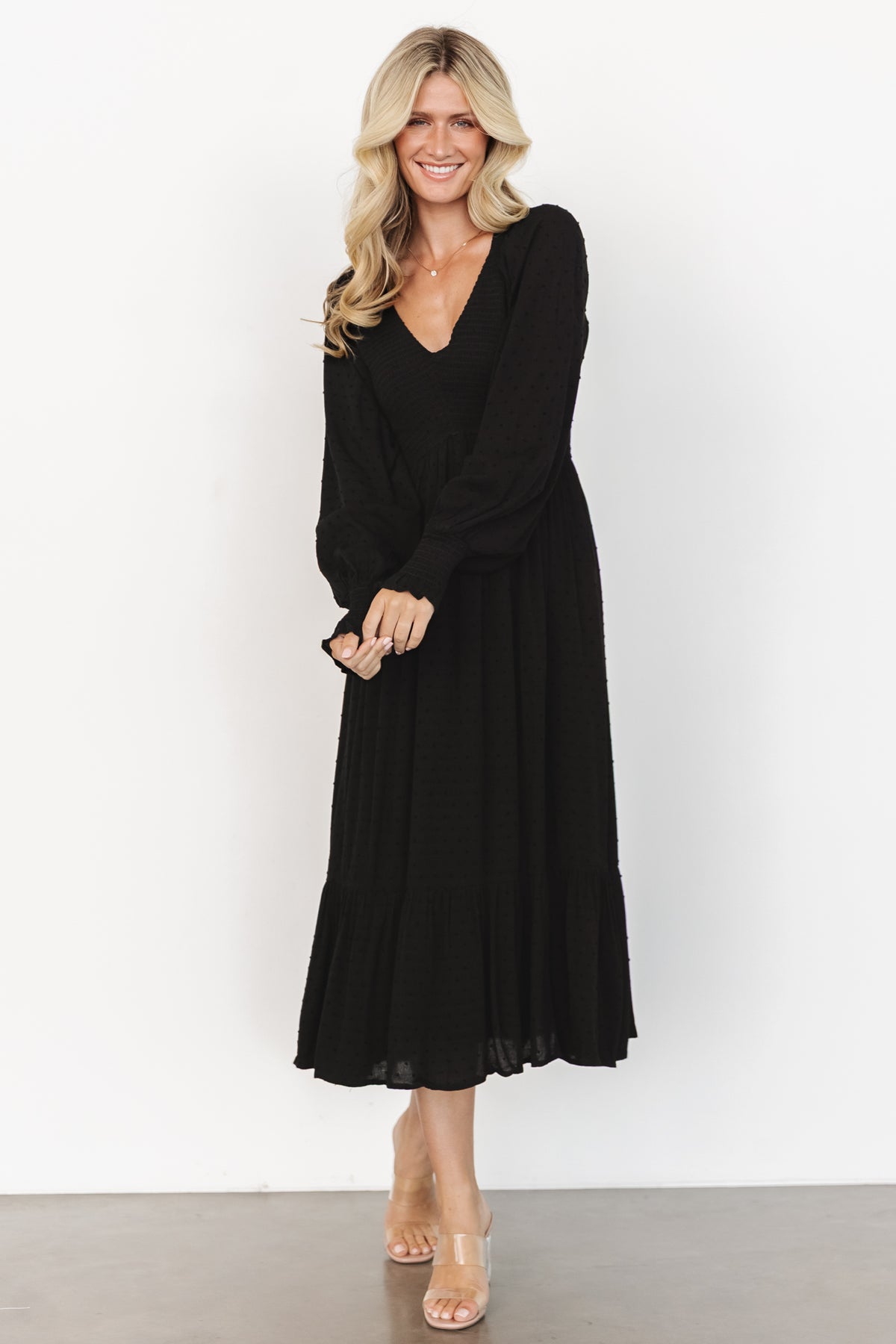 Full-length view of black Lainey smocked dotted midi dress on model, highlighting silhouette, V-neckline, tiered hem, and easy styling for day or evening events.