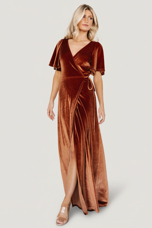 Front view of Meghan Velvet Wrap Maxi Dress in spice; model standing, V-neck and waist tie visible, full-length tulip hem and drape.
