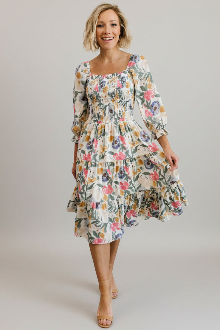 Front view of a smocked midi dress in muted multi floral print on a model, showing square neckline and tiered skirt, full-length.