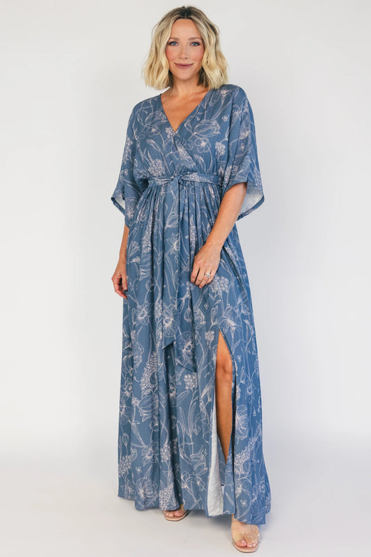 Model wearing a navy botanical-print maxi dress with a surplice V-neckline, kimono sleeves, an elastic waist with a removable sash tie, and a flowing full-length skirt featuring a side slit.