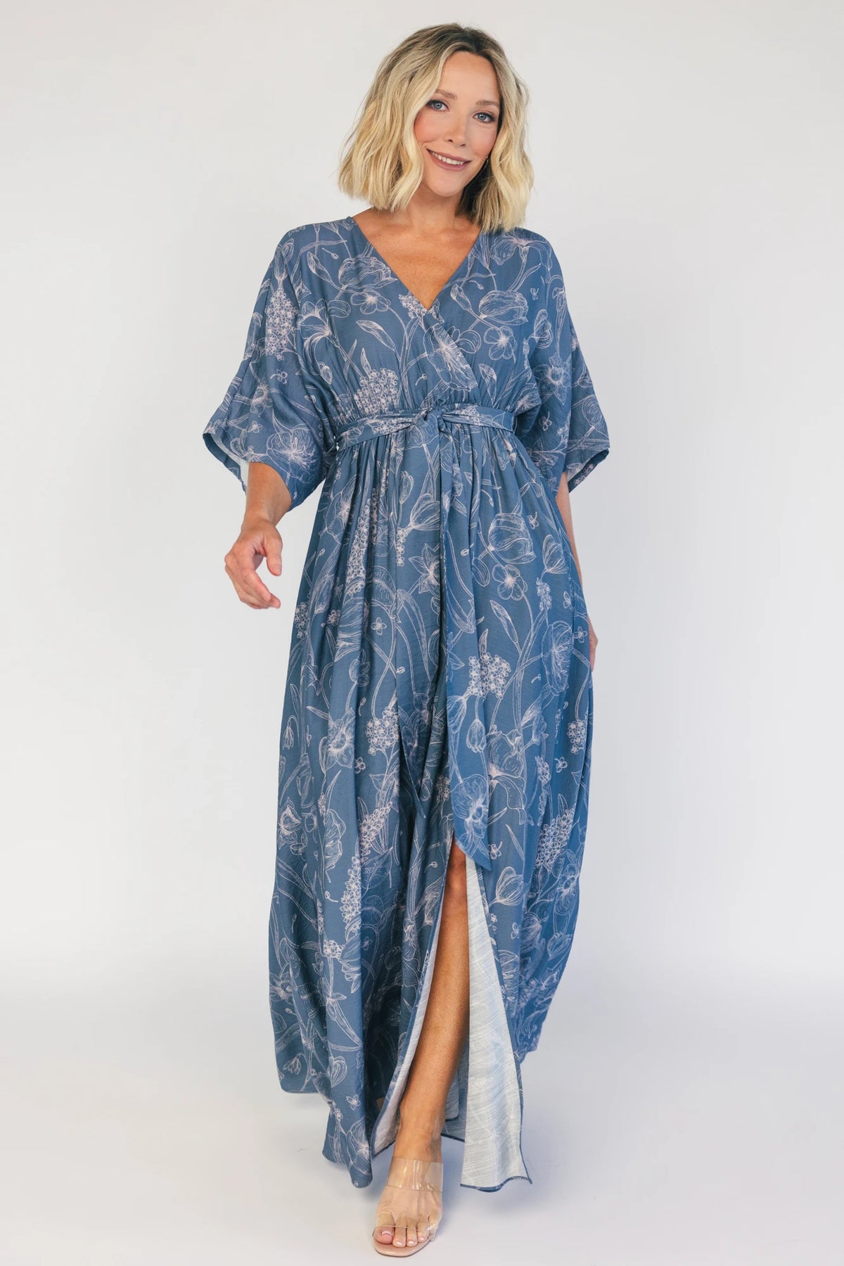 Model wearing a navy botanical-print maxi dress with a surplice V-neckline, kimono sleeves, an elastic waist with a removable sash tie, and a flowing full-length skirt featuring a side slit.