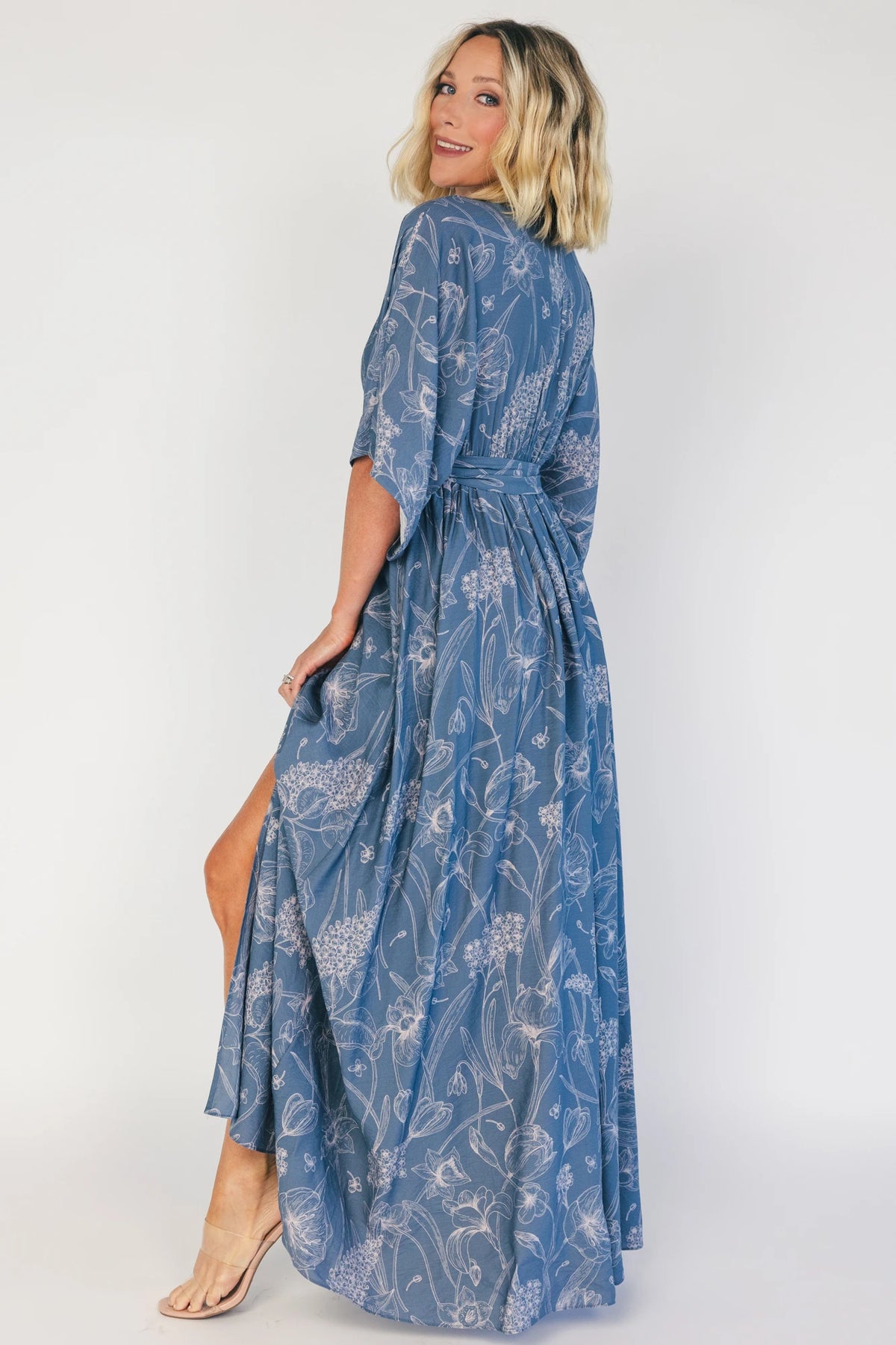 Model wearing a navy botanical-print maxi dress with a surplice V-neckline, kimono sleeves, an elastic waist with a removable sash tie, and a flowing full-length skirt featuring a side slit.