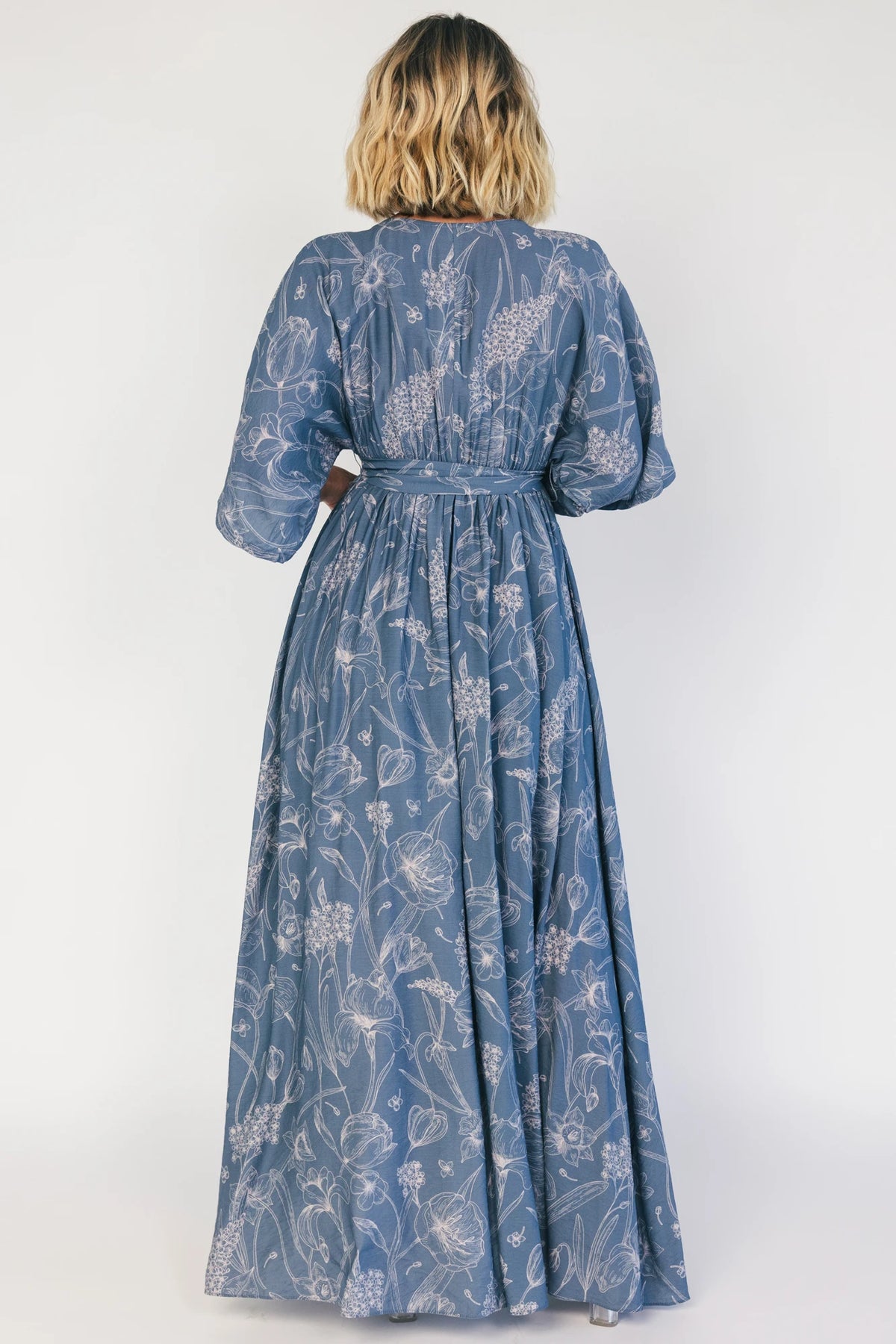 Model wearing a navy botanical-print maxi dress with a surplice V-neckline, kimono sleeves, an elastic waist with a removable sash tie, and a flowing full-length skirt featuring a side slit.