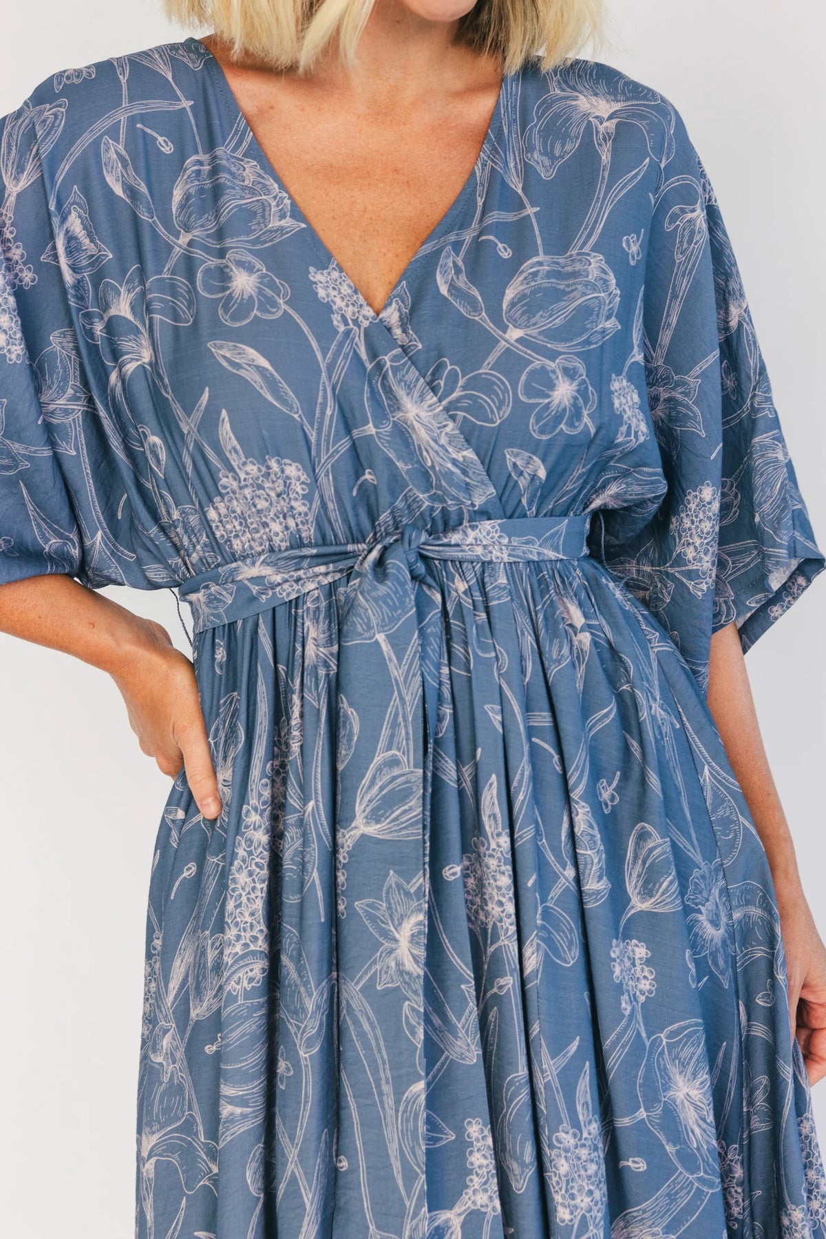 Model wearing a navy botanical-print maxi dress with a surplice V-neckline, kimono sleeves, an elastic waist with a removable sash tie, and a flowing full-length skirt featuring a side slit.