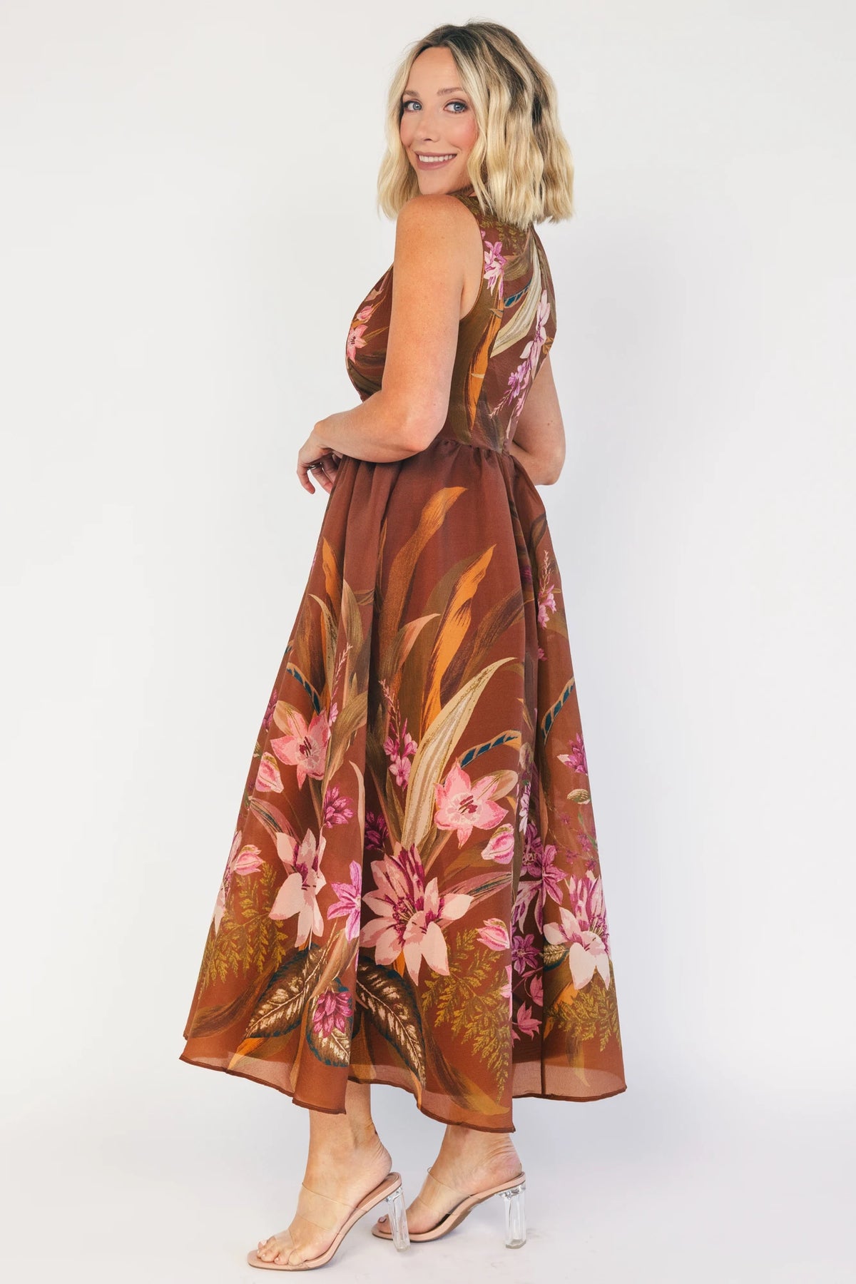 Model wearing a brown floral midi dress with a high boat neckline, sleeveless fitted bodice, and defined waist. The dress features a pink floral print throughout and a flowing midi-length skirt, creating an elegant, timeless silhouette.