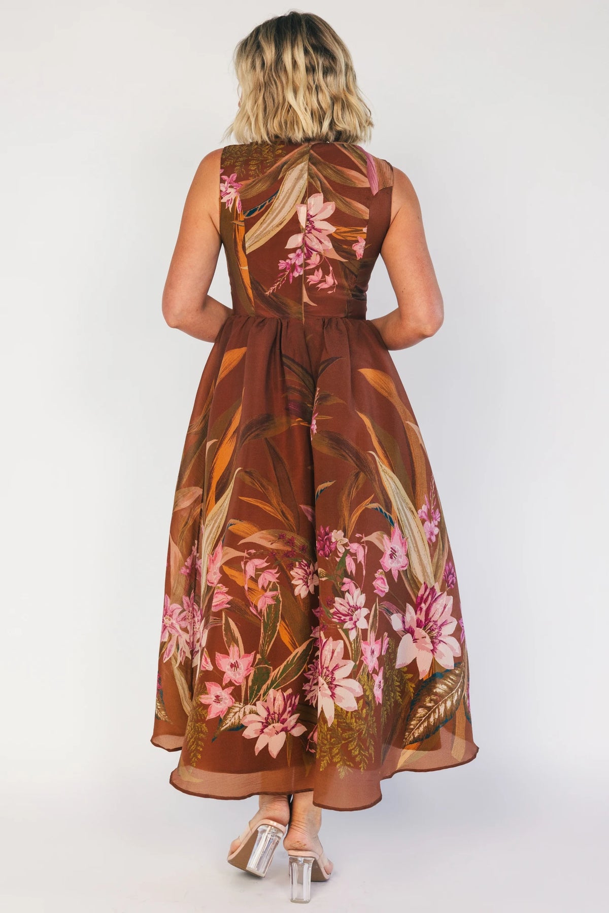 Model wearing a brown floral midi dress with a high boat neckline, sleeveless fitted bodice, and defined waist. The dress features a pink floral print throughout and a flowing midi-length skirt, creating an elegant, timeless silhouette.