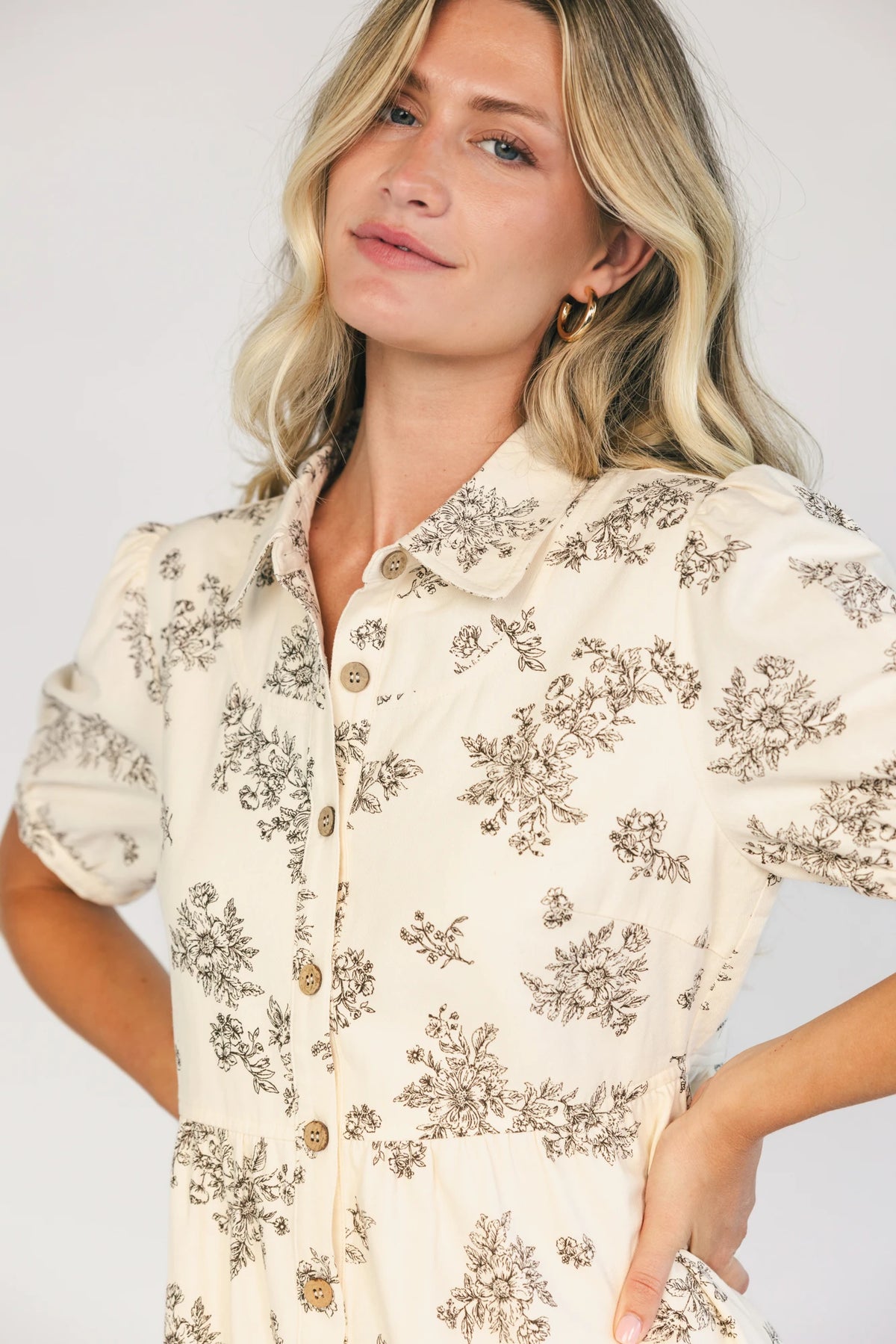 Model wearing a cream floral short dress with a collared neckline, button-front closure, short gathered sleeves, and a relaxed empire-waist silhouette.