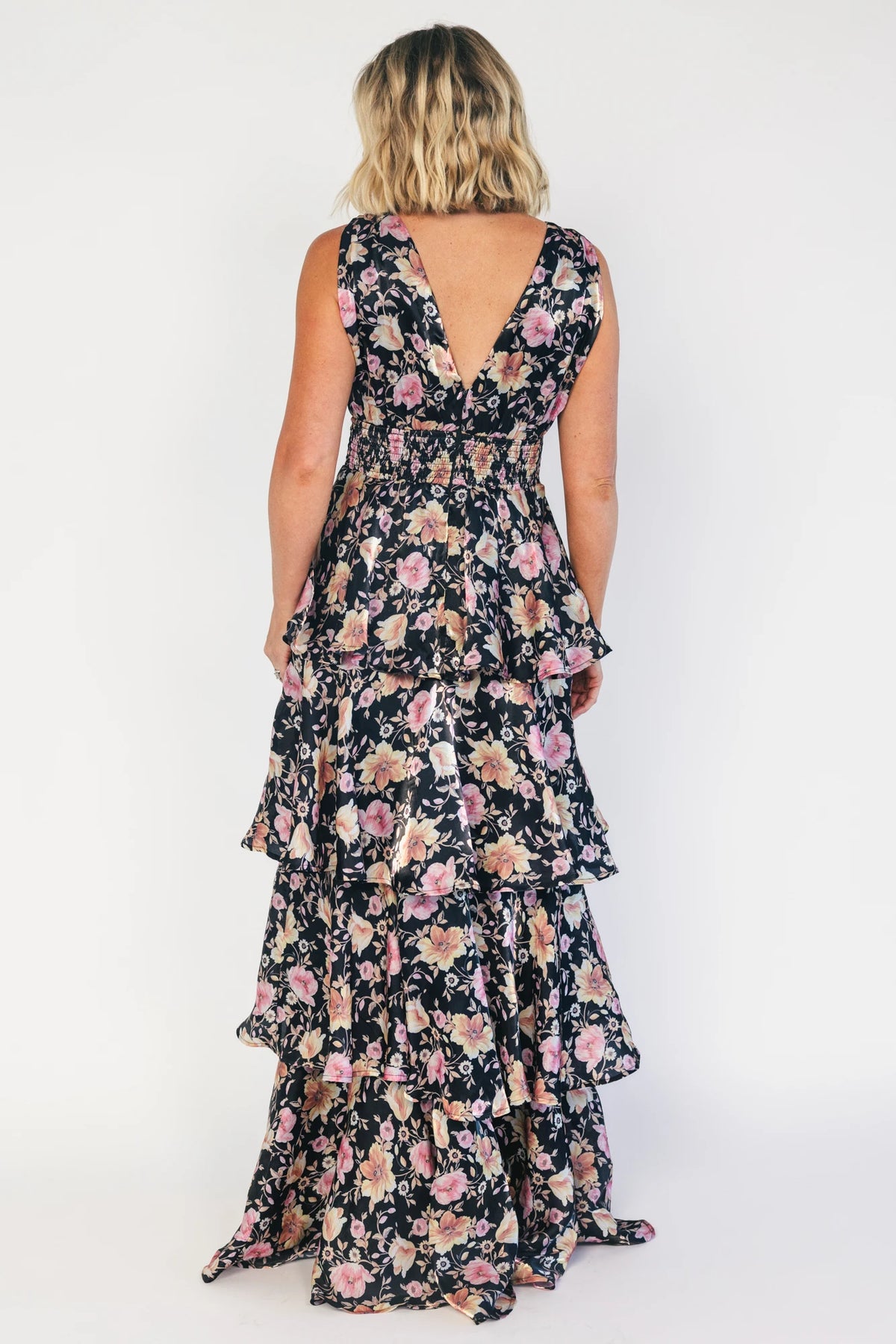 Model wearing a black floral maxi dress with a deep V-neckline, banded waist, and cascading tiered skirt. The dress features a black background with pink, yellow, and orange floral print, creating a flowing, romantic silhouette.
