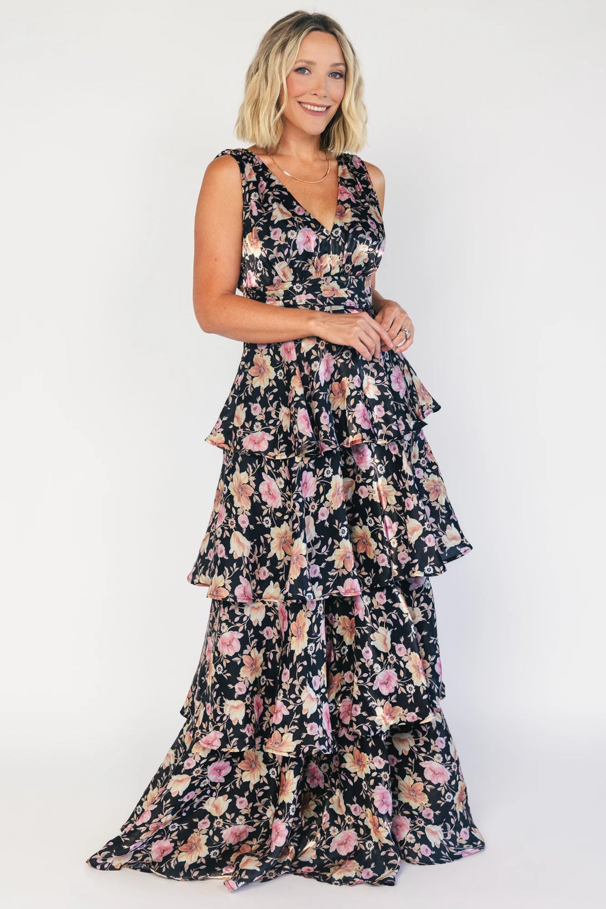 Model wearing a black floral maxi dress with a deep V-neckline, banded waist, and cascading tiered skirt. The dress features a black background with pink, yellow, and orange floral print, creating a flowing, romantic silhouette.