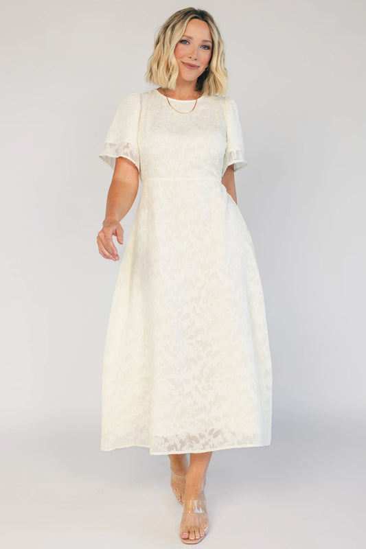 Model wearing a cream midi dress with a round neckline, short gathered sleeves, and a fitted bodice.