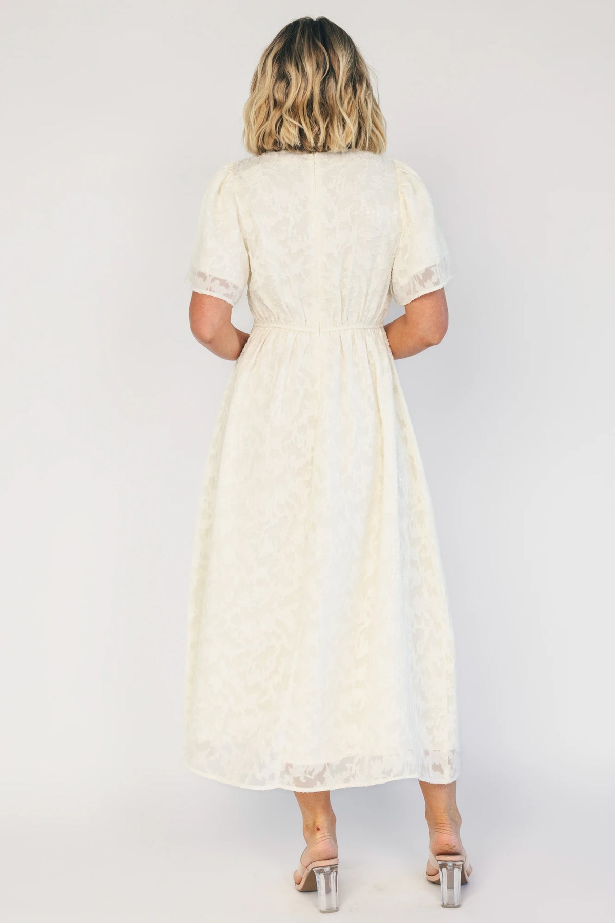 Model wearing a cream midi dress with a round neckline, short gathered sleeves, and a fitted bodice.