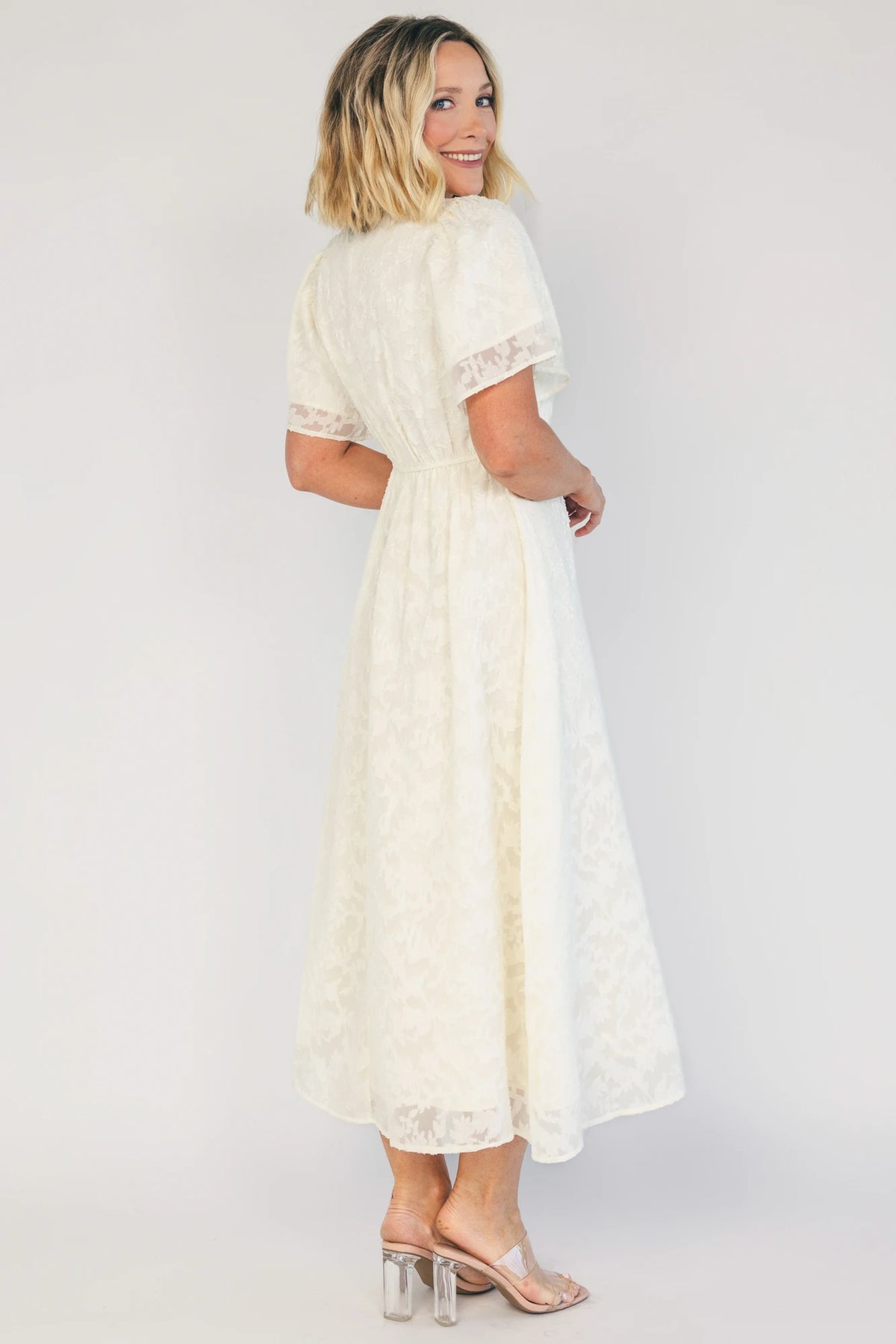 Model wearing a cream midi dress with a round neckline, short gathered sleeves, and a fitted bodice.