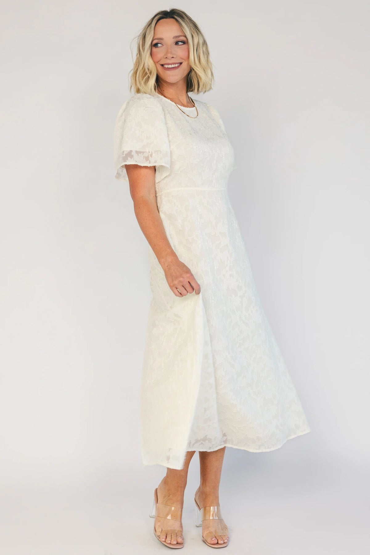 Model wearing a cream midi dress with a round neckline, short gathered sleeves, and a fitted bodice.