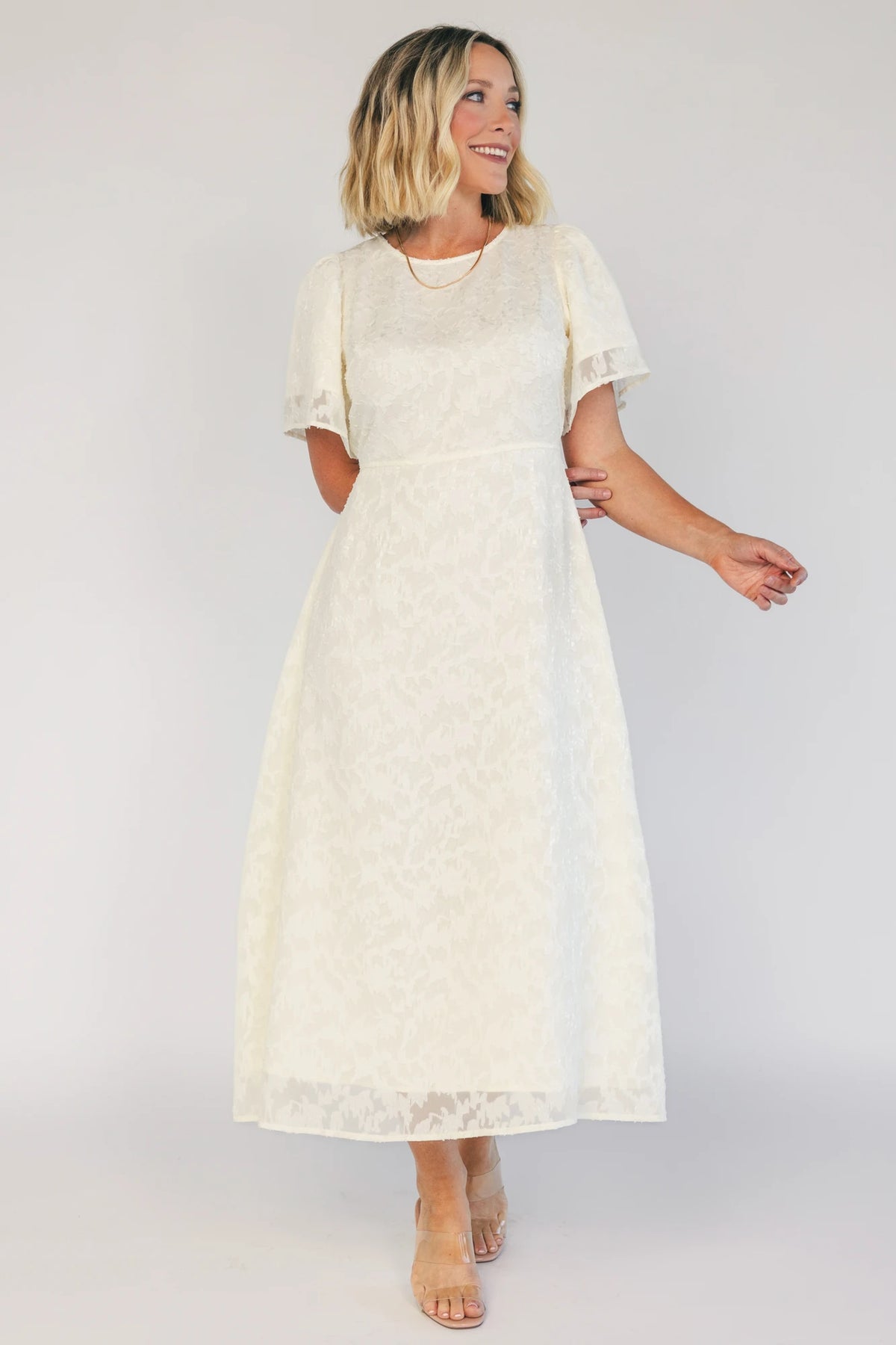Model wearing a cream midi dress with a round neckline, short gathered sleeves, and a fitted bodice.