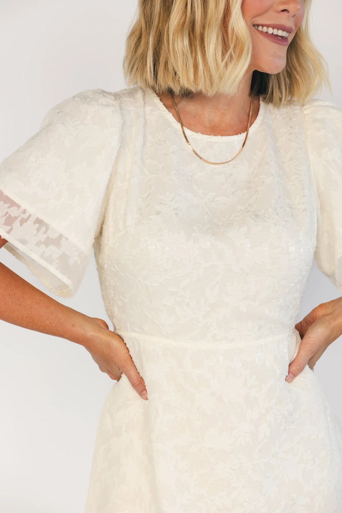 Model wearing a cream midi dress with a round neckline, short gathered sleeves, and a fitted bodice.