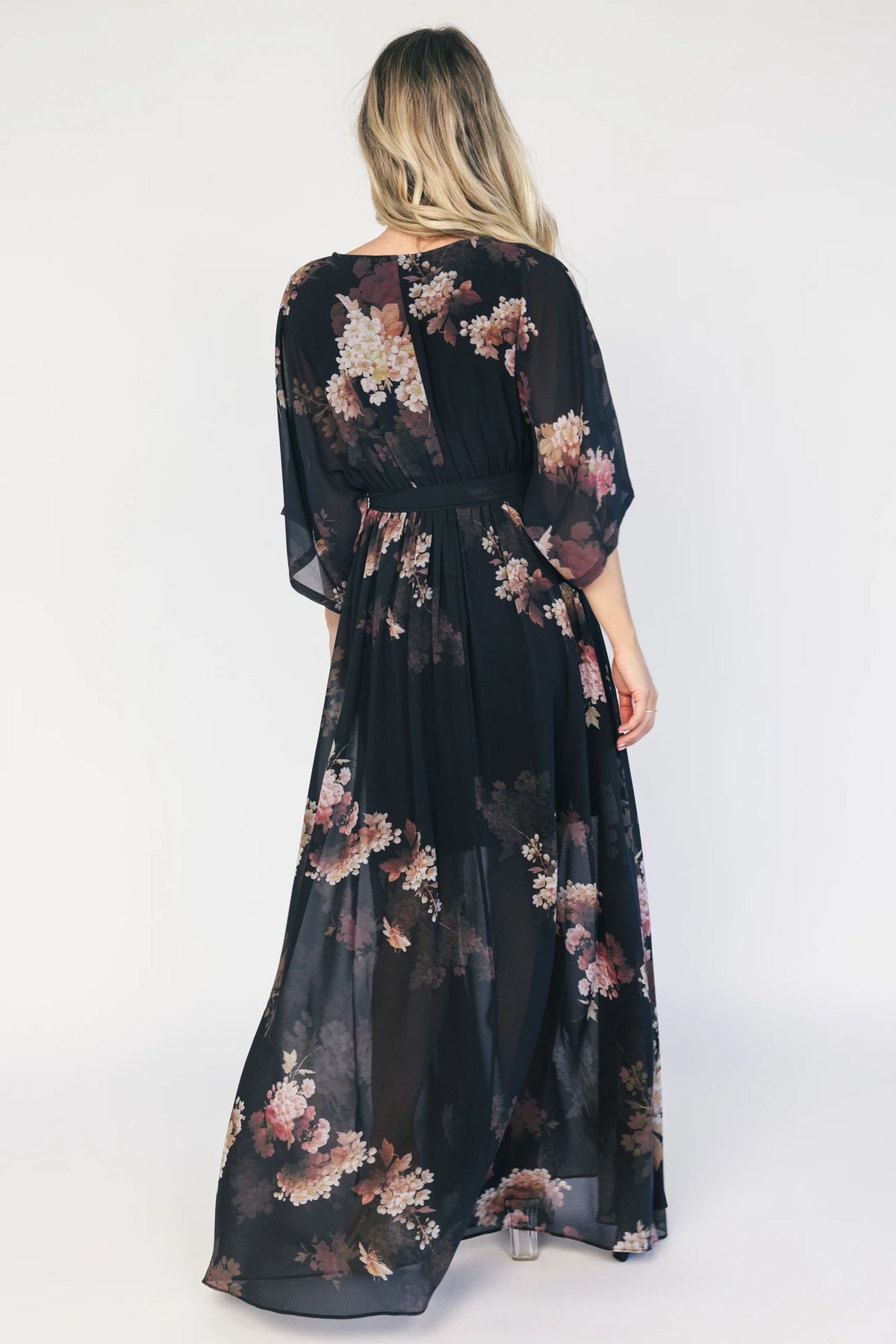 Model wearing a black floral maxi dress with a surplice V-neckline, kimono sleeves, and an elastic waist with a removable sash tie.