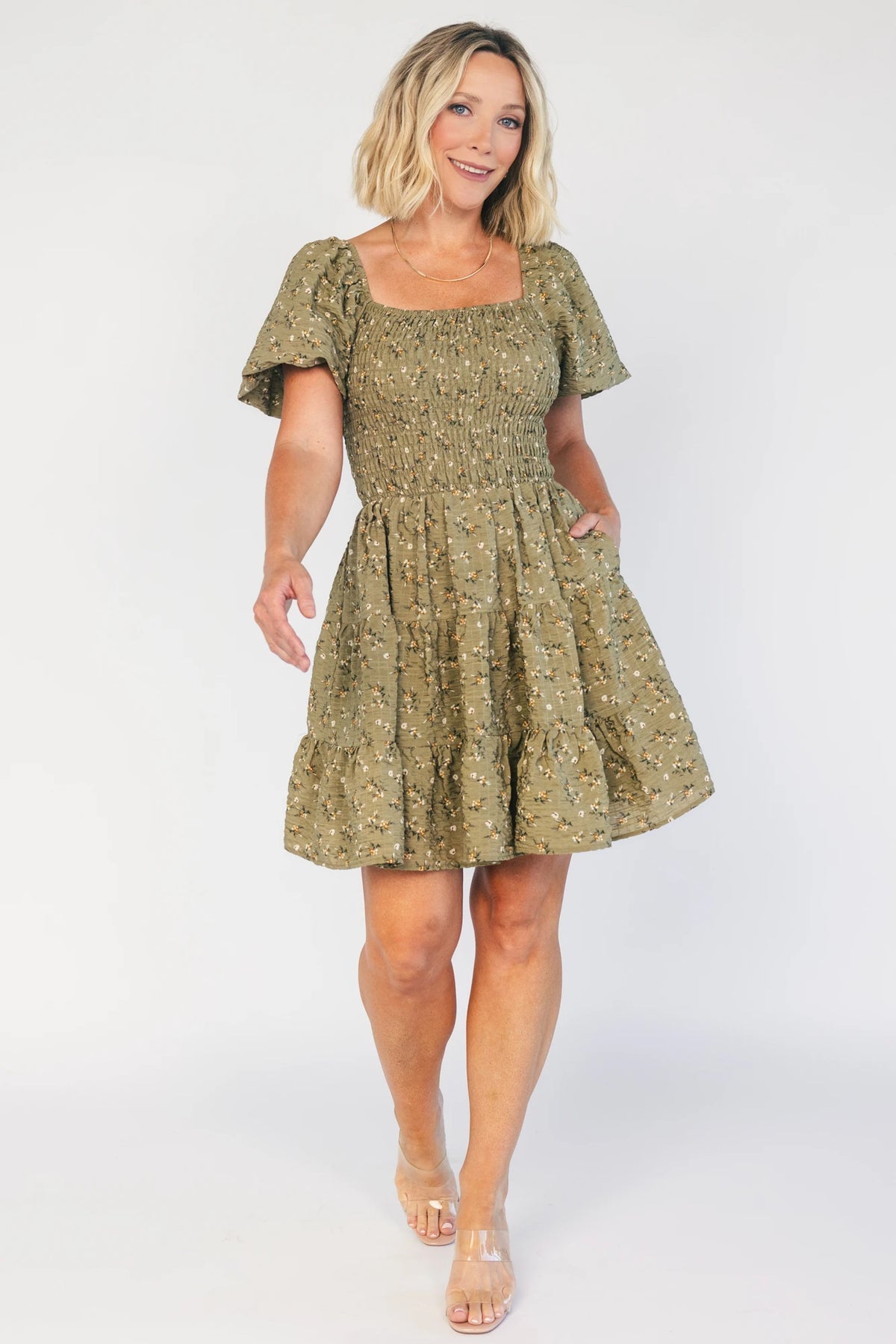 Model wearing an olive floral short dress with a square neckline, fully smocked bodice, and short balloon sleeves.