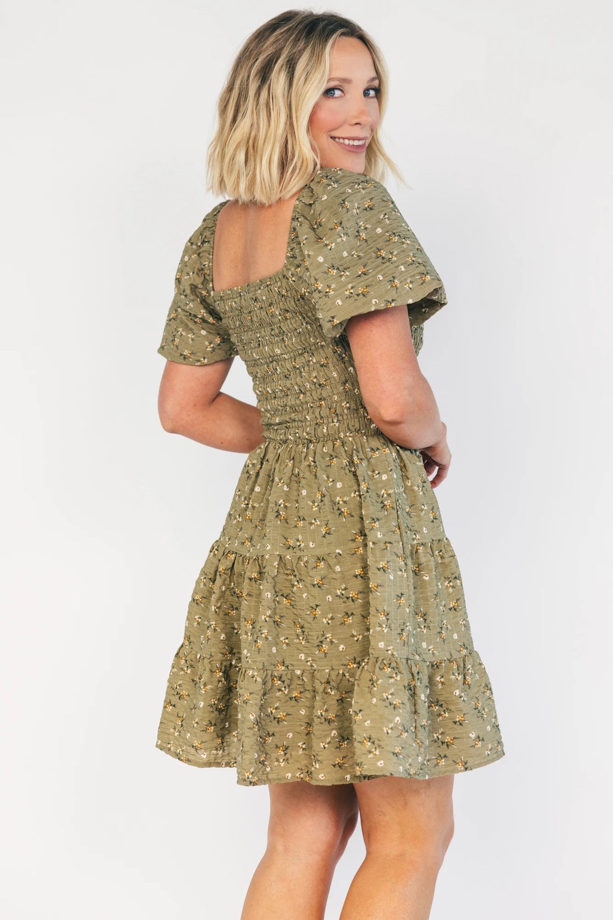 Model wearing an olive floral short dress with a square neckline, fully smocked bodice, and short balloon sleeves.