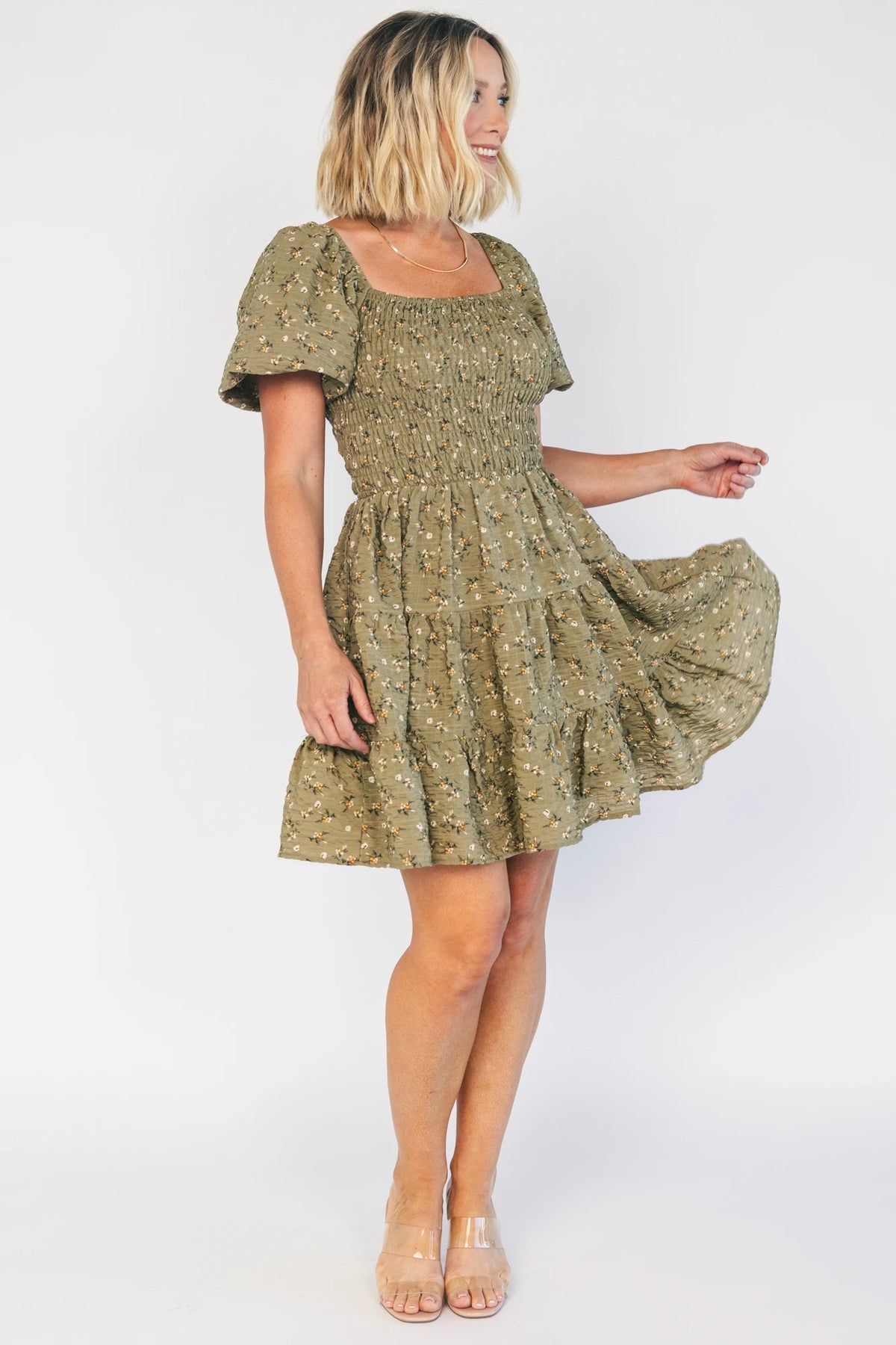 Model wearing an olive floral short dress with a square neckline, fully smocked bodice, and short balloon sleeves.