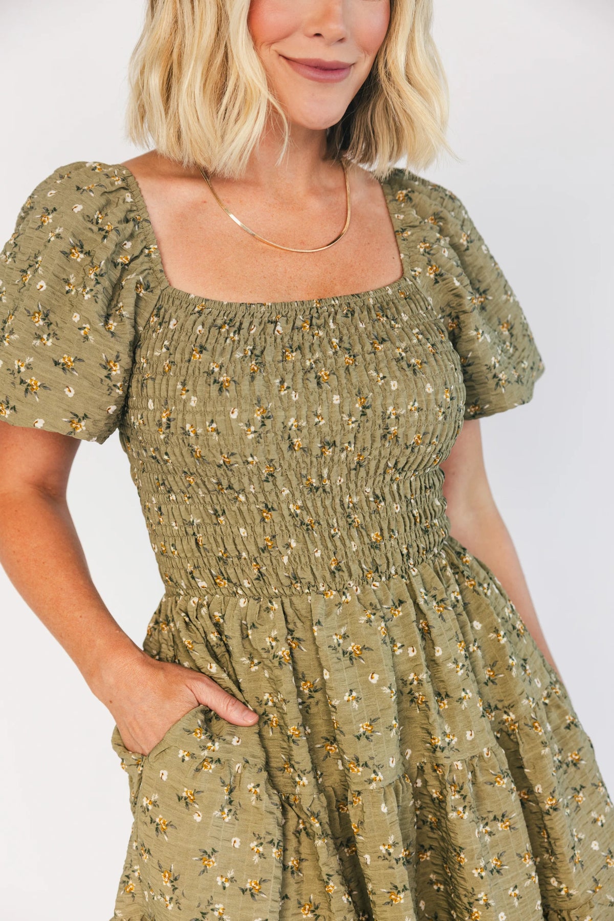 Model wearing an olive floral short dress with a square neckline, fully smocked bodice, and short balloon sleeves.