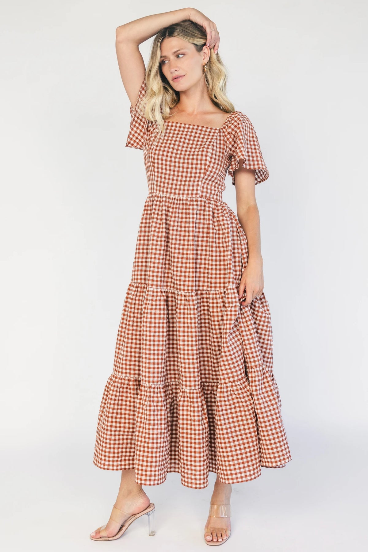 Model wearing a spice and white gingham maxi dress with a square neckline, flutter sleeves, and a fitted bodice with a smocked back