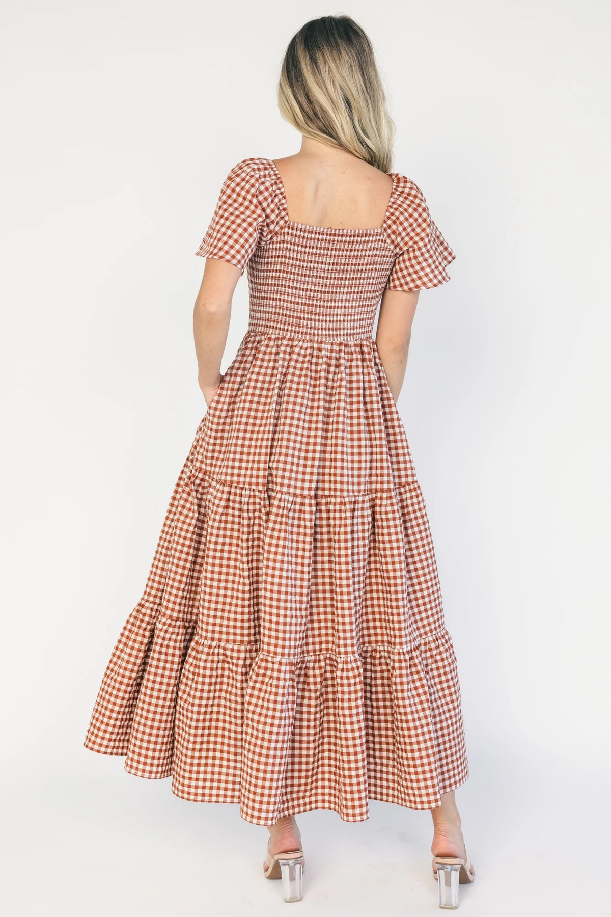 Model wearing a spice and white gingham maxi dress with a square neckline, flutter sleeves, and a fitted bodice with a smocked back