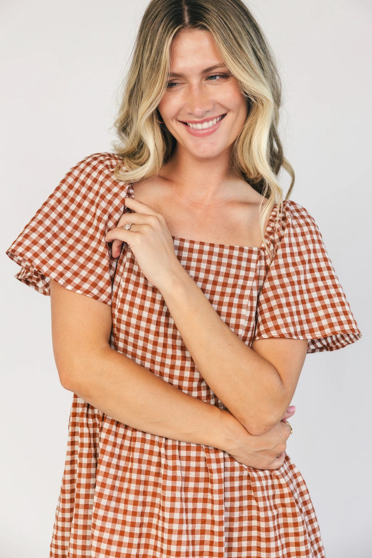 Model wearing a spice and white gingham maxi dress with a square neckline, flutter sleeves, and a fitted bodice with a smocked back