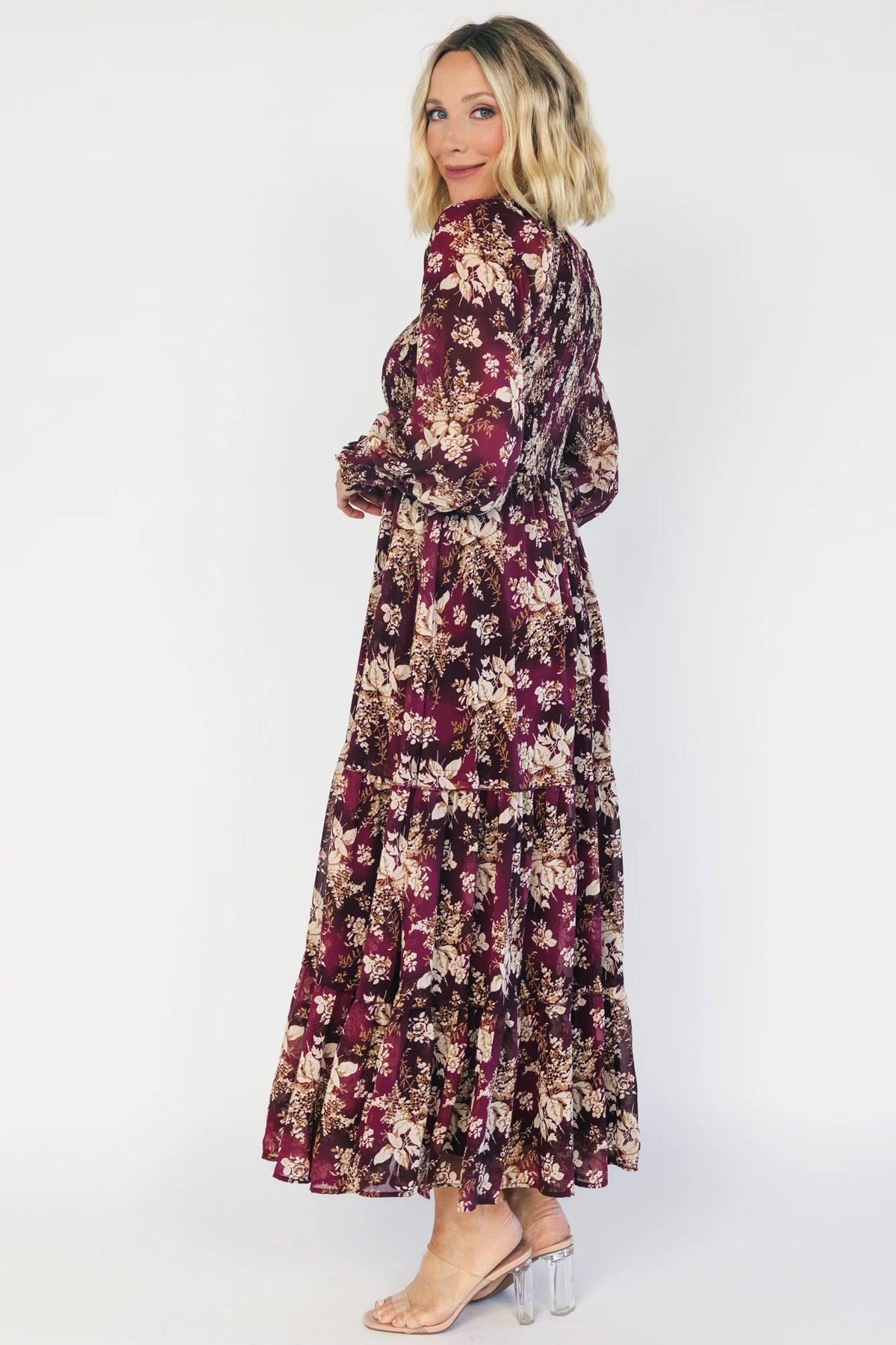Model wearing a burgundy floral maxi dress with a V-neckline, smocked bodice, and long sheer sleeves with smocked ruffle cuffs.
