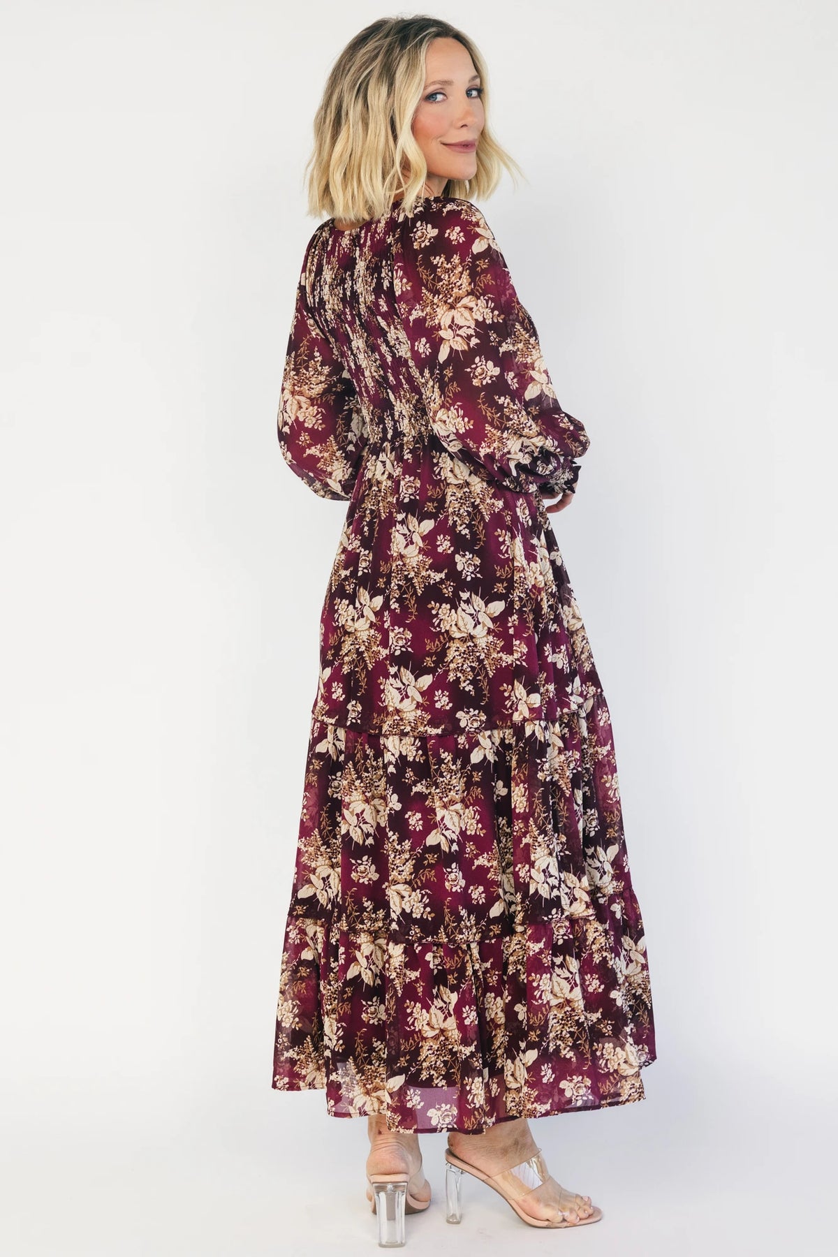 Model wearing a burgundy floral maxi dress with a V-neckline, smocked bodice, and long sheer sleeves with smocked ruffle cuffs.